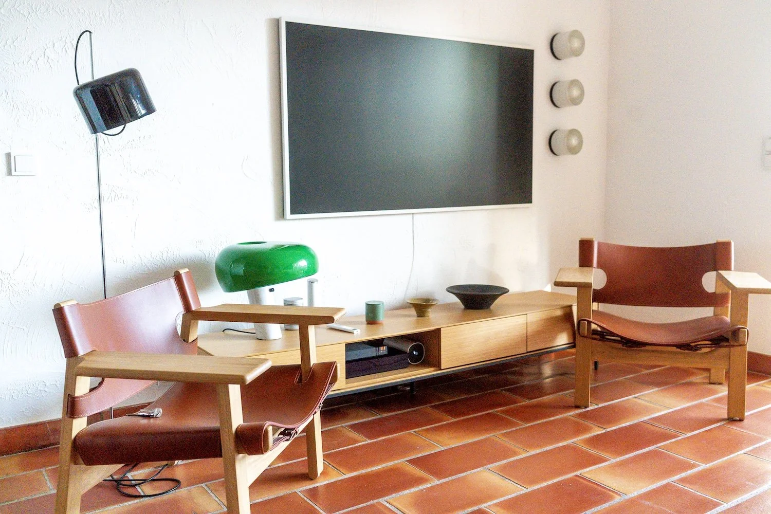 Living room with two wooden chairs with reddish-brown leather cushions, a wooden TV console, a large flat-screen TV on the wall, and decorative objects including a green table lamp and ceramic bowls. The floor is tiled with reddish-brown tiles.