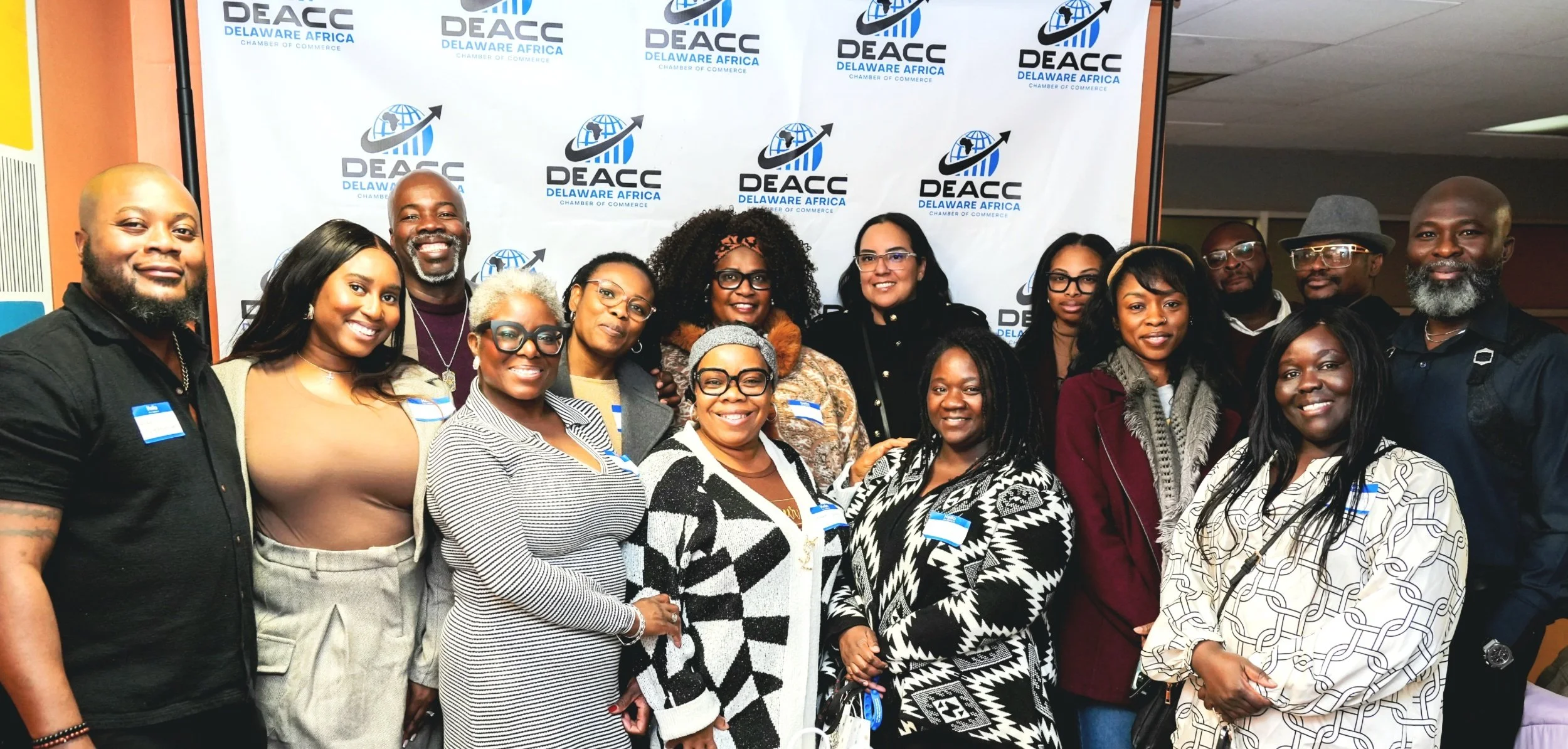 A group of diverse men and women smiling and posing together in front of a backdrop with the DEACC Delaware Africa logo at an event.