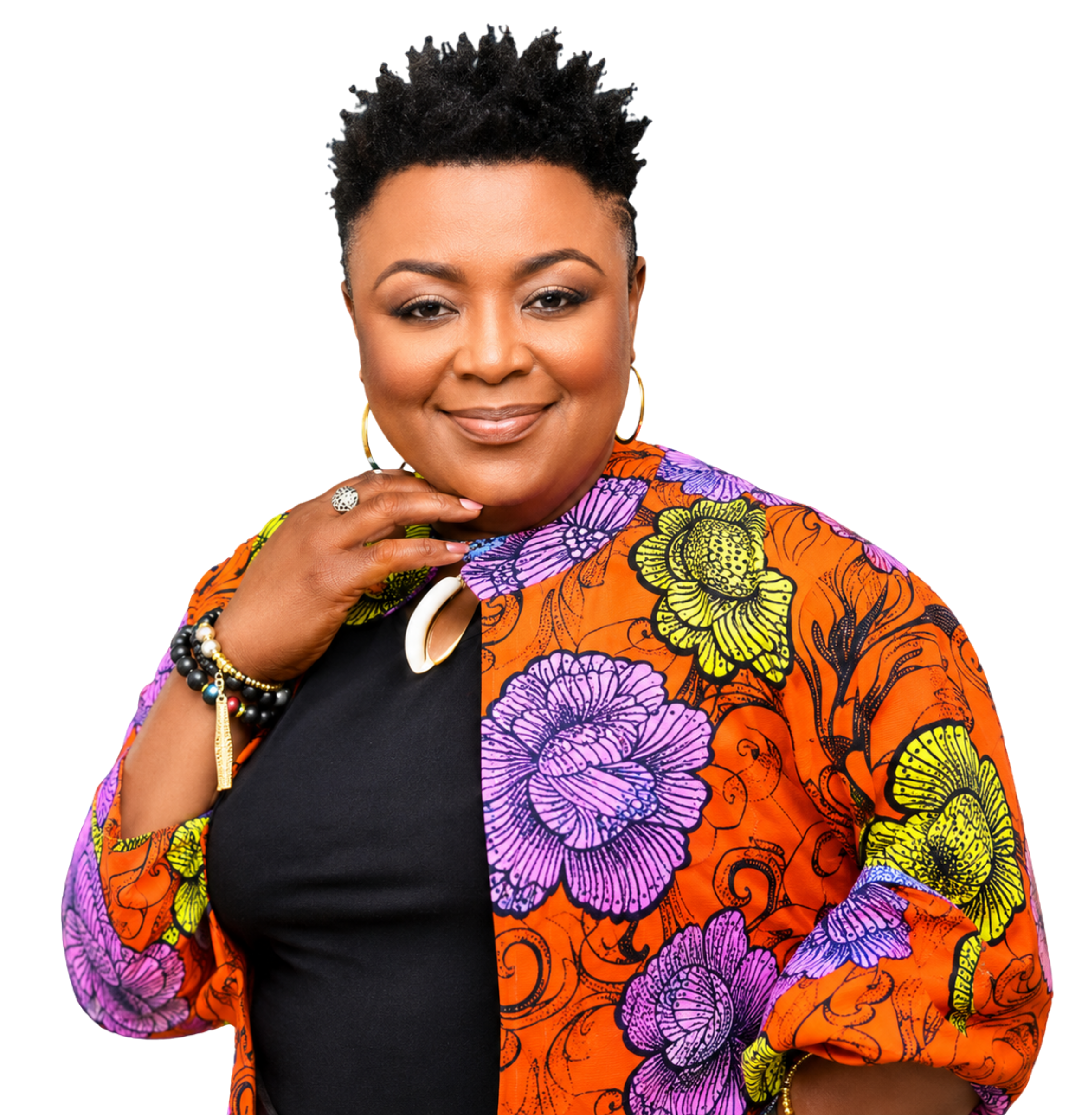 A woman with short curly hair, wearing a colorful floral patterned jacket over a black shirt, accessorized with bracelets, earrings, and a ring, smiling and looking directly at the camera.