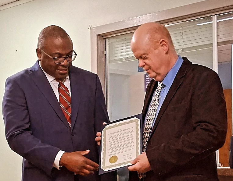 Two men in business suits, one presenting a framed certificate to the other, indoors with a window in the background.