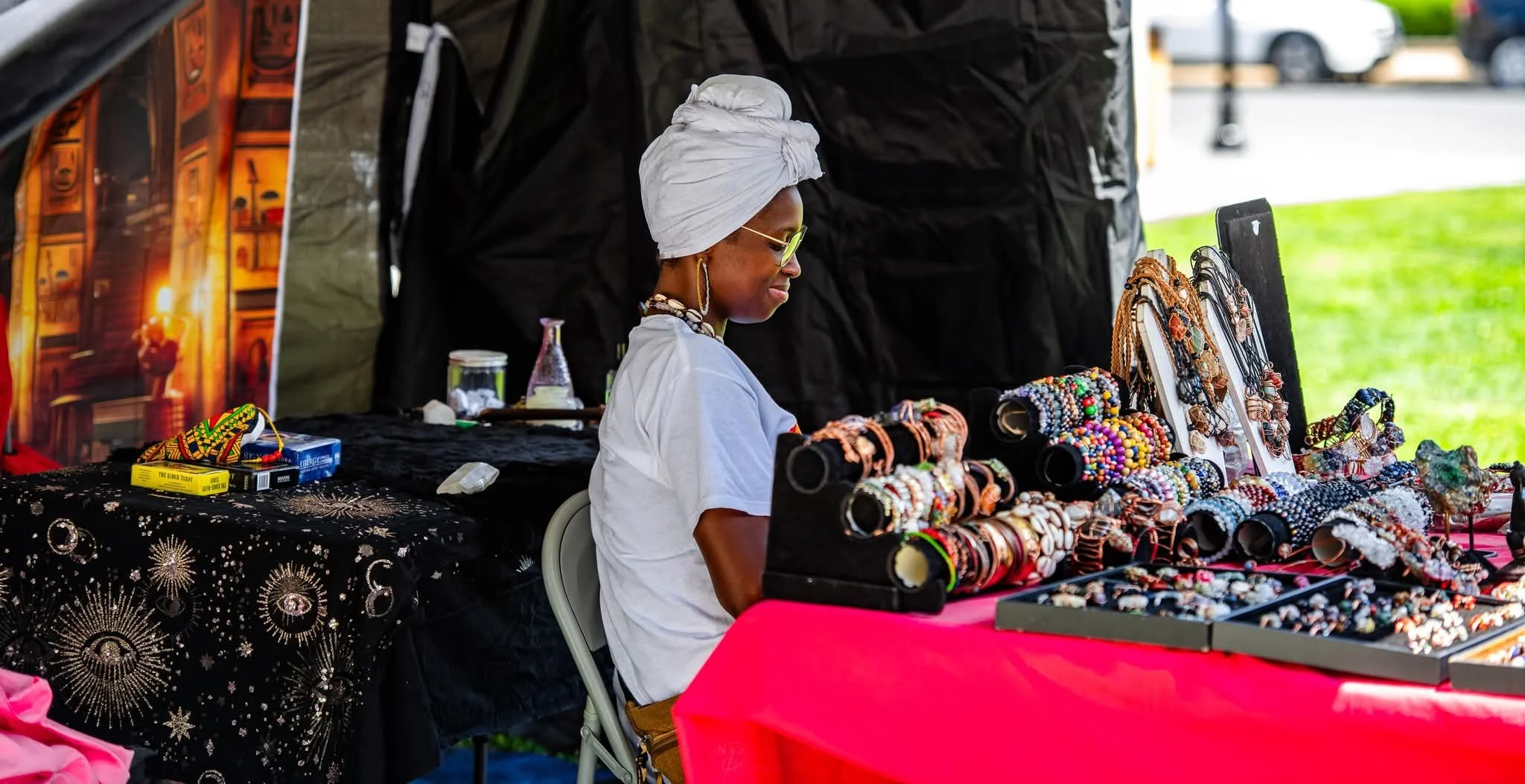 A woman wearing sunglasses, a white head wrap, and white clothing sits at a vendor stall displaying colorful jewelry, including bracelets and necklaces, under a black canopy on a sunny day.