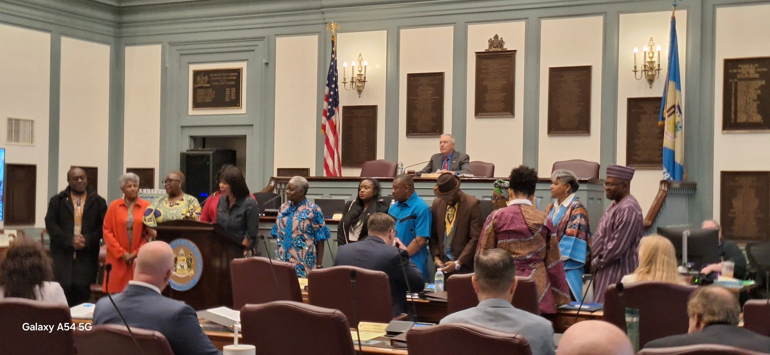 A diverse group of people standing in front of a speaker at a formal event or government meeting in a large room with dark wood paneling, flags, and plaques on the wall.