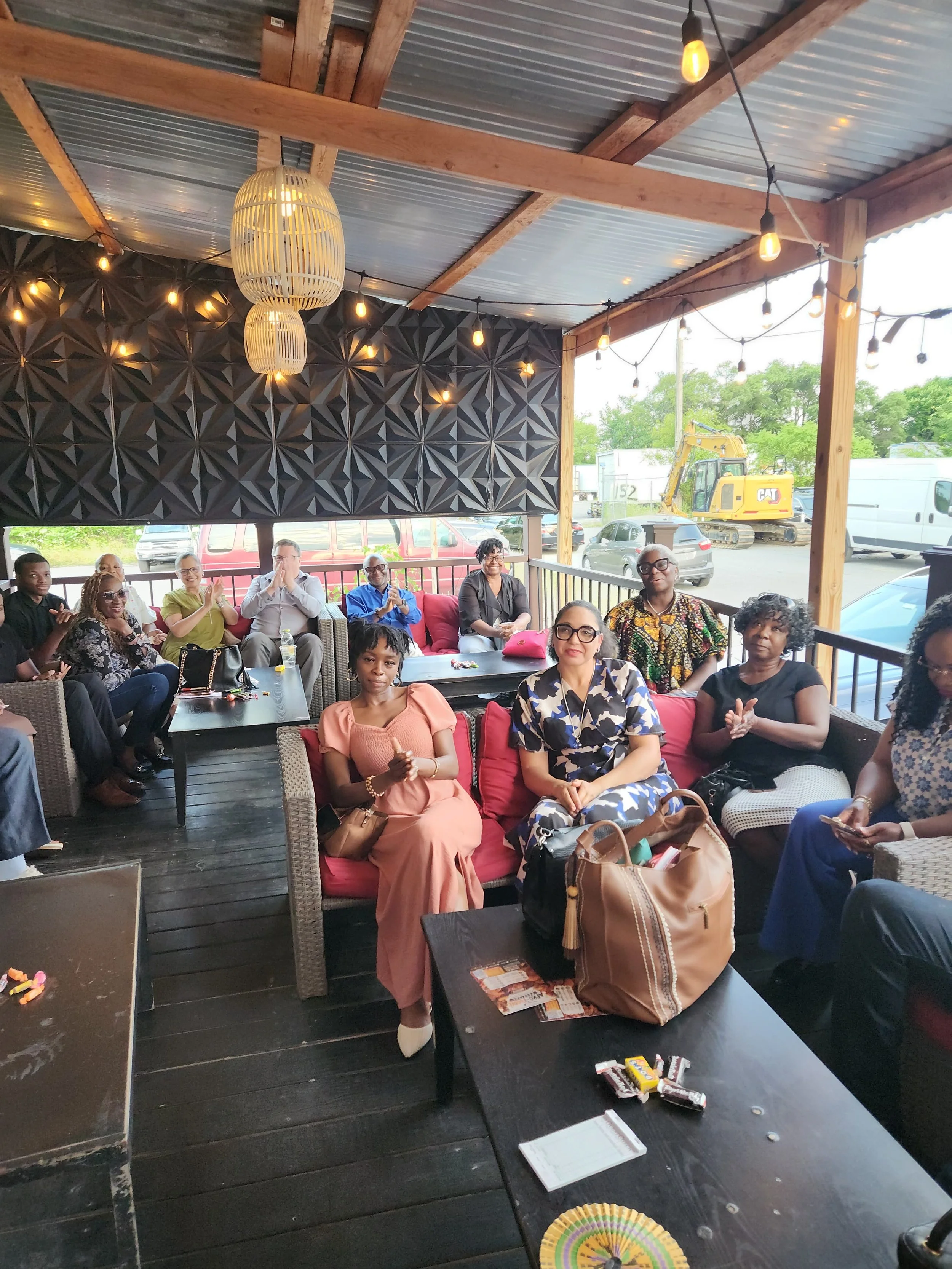 A group of people sitting on outdoor patio furniture at a gathering, with some clapping and others looking at the camera. The patio has a wooden ceiling with hanging lightbulbs and decorative lanterns, and a view of parked cars and construction equipment outside.