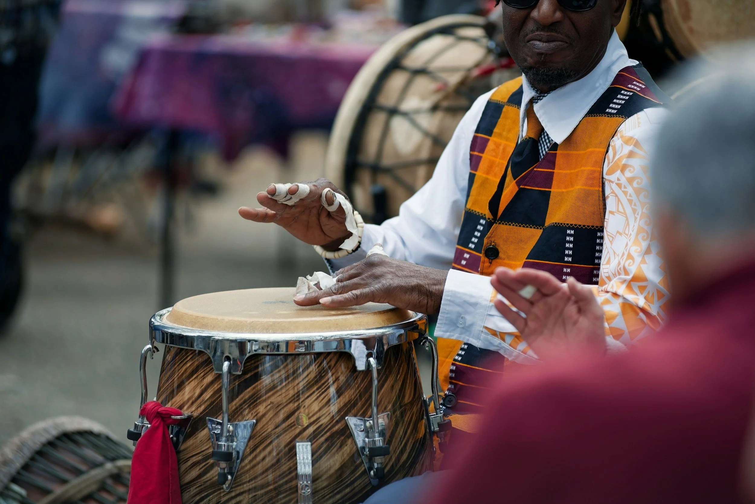 A man wearing sunglasses and a patterned shirt is playing a drum with his hands at an outdoor event. The drum has a wooden finish and is decorated with a red cloth tied around it. Other drums and blurred figures are visible in the background.