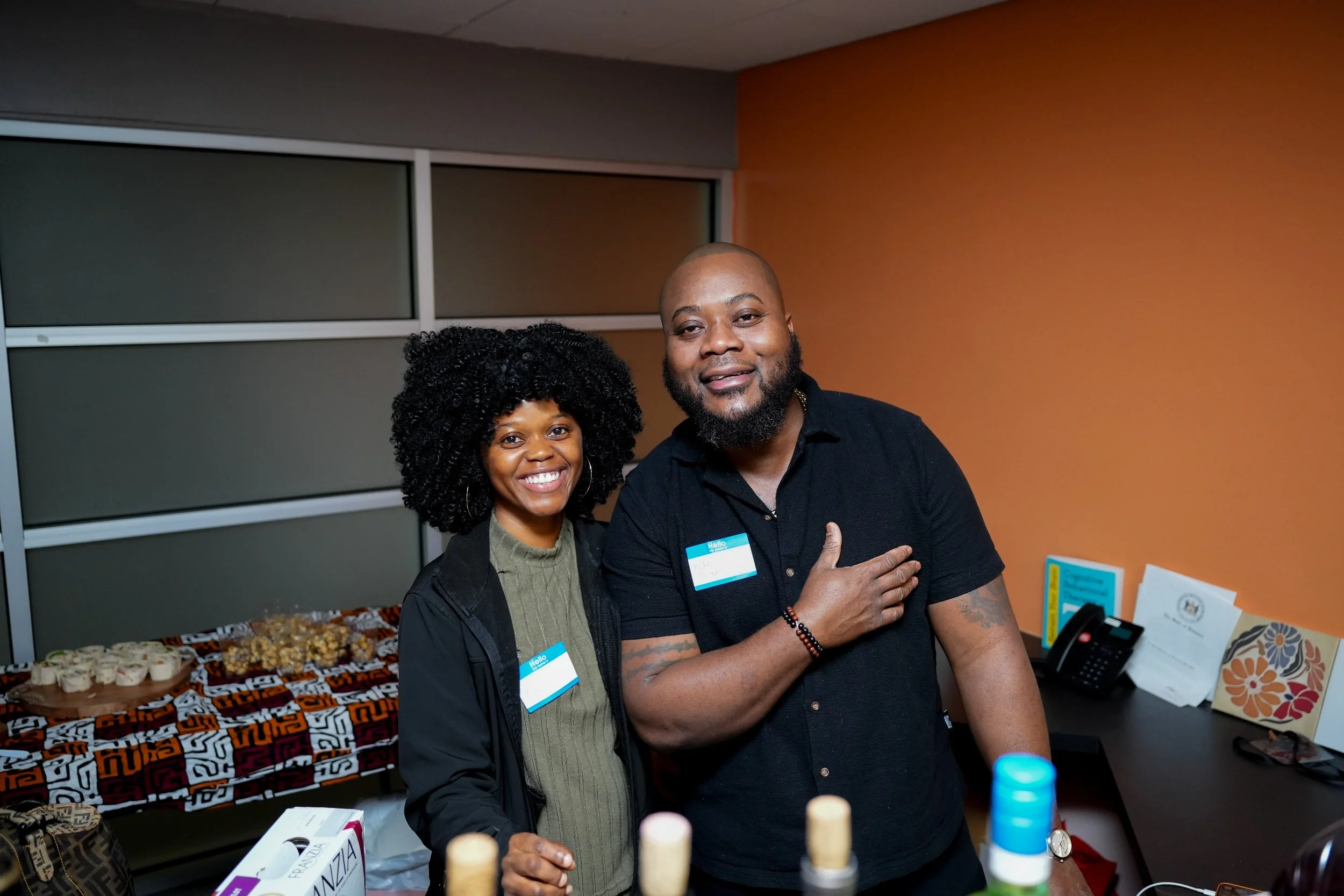 A woman and man smiling at a social event, standing near a table with food and drinks, in an indoor setting with orange and gray walls.