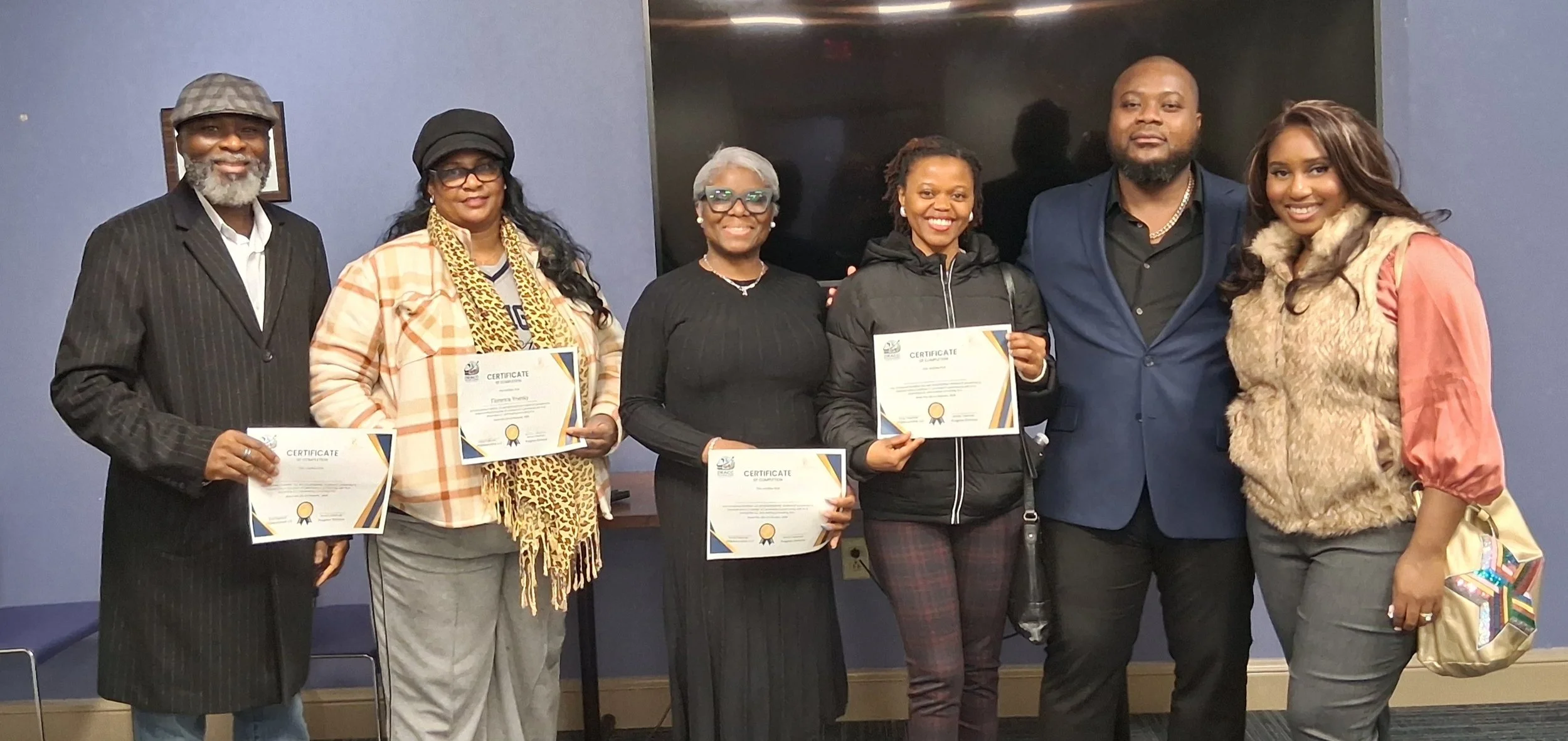 Group of six diverse adults posing for a photo, some holding certificates, in an indoor setting with a blue-gray wall and a large black screen behind them.