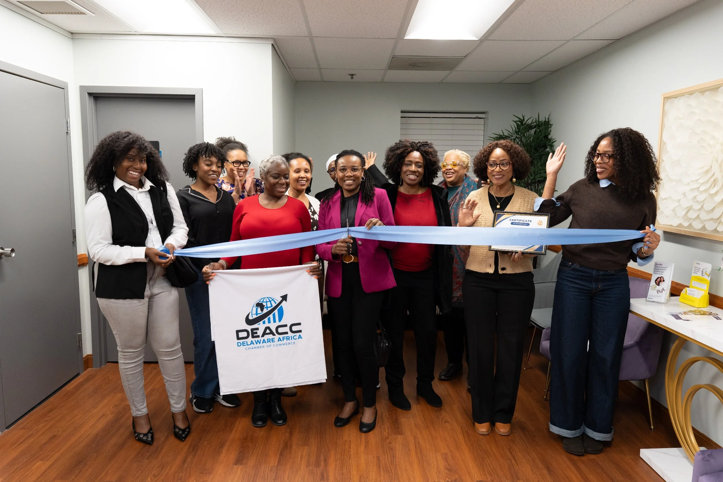 Group of women gathered indoors, holding a ribbon cuttings and signage for DEACC Deaware Africa Chamber of Commerce, smiling and celebrating an event.
