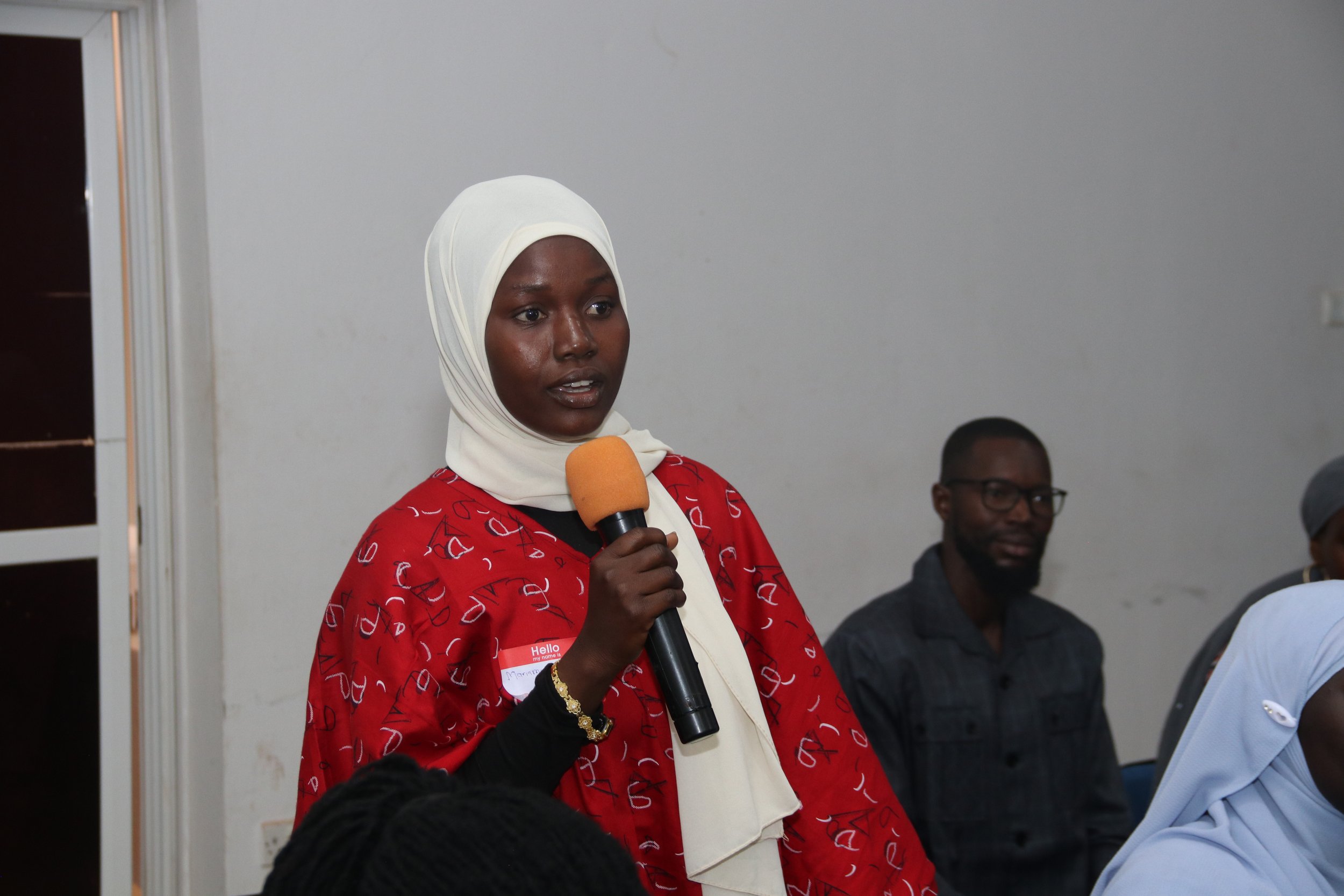 A woman with a white headscarf and a red patterned jacket is speaking into a microphone in a room with other seated people, including a man with glasses.