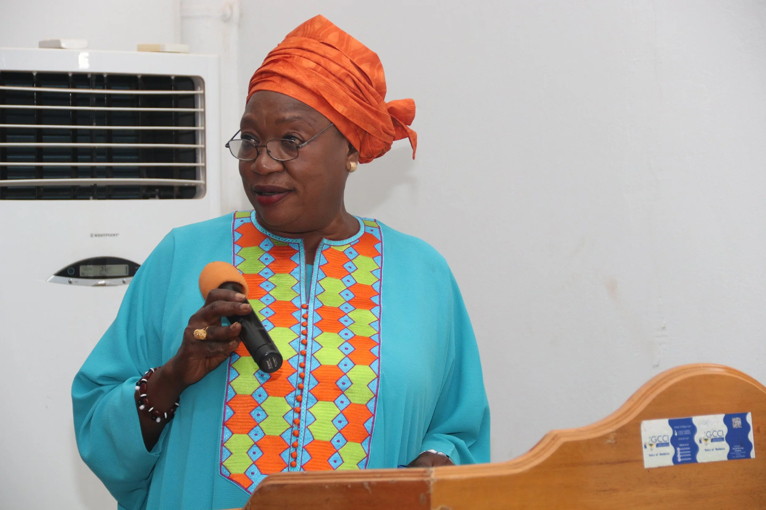 Woman wearing glasses, orange headwrap, and colorful embroidered dress, speaking into a microphone at a podium. An air conditioning unit is visible in the background.