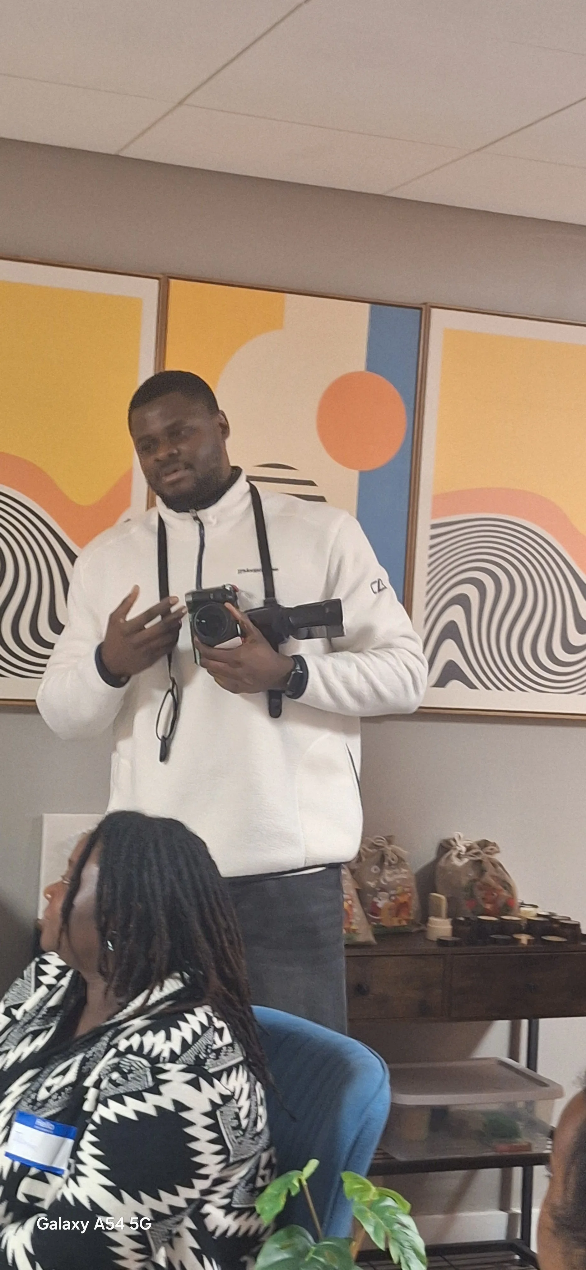 A man wearing a white jacket holding a camera and pointing to himself. He appears to be talking or explaining something during a gathering or event with colorful abstract wall art in the background.