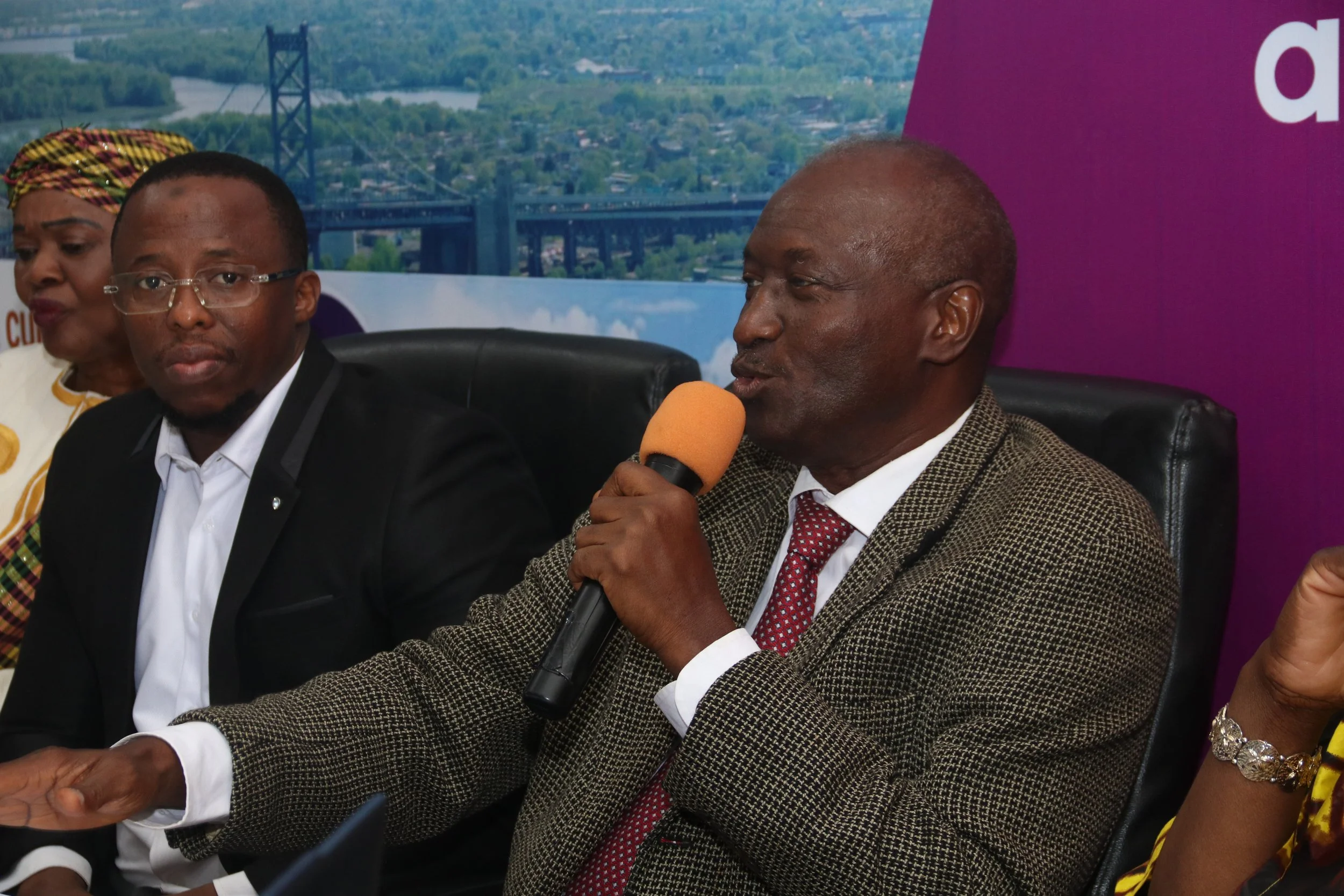 A man in a suit and red tie speaking into a microphone during a panel discussion, seated next to a woman with a colorful headwrap and a woman in a white coat, with a cityscape and bridge background.
