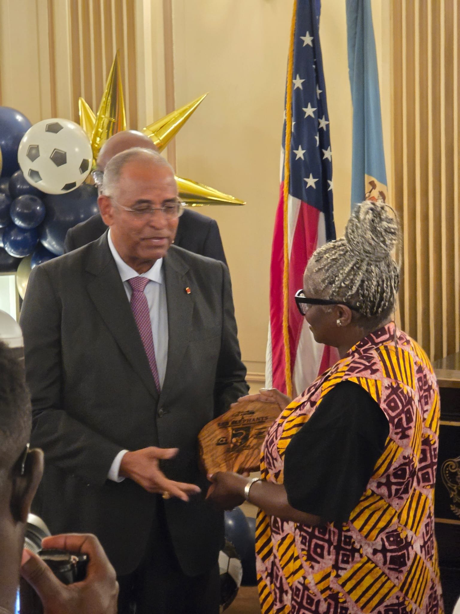 A man in a suit and a woman in a colorful patterned dress shaking hands during a formal event. There are American and another flag visible in the background, along with balloons and a large gold star decoration.