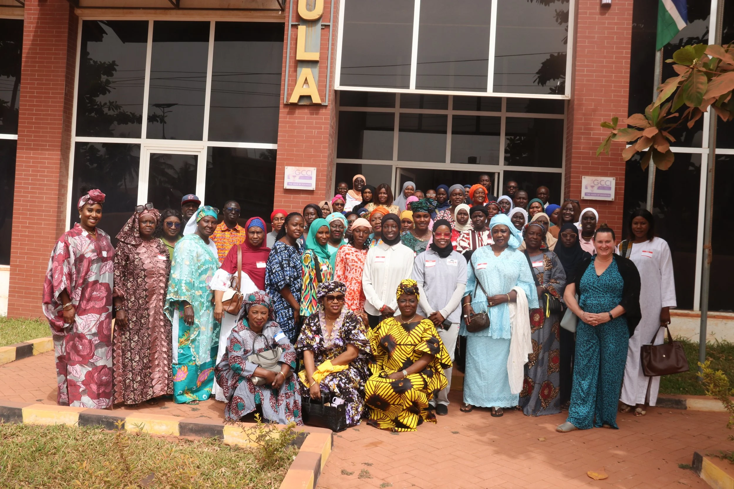 A large group of women and a few men standing outside a modern brick building with glass windows, group photo, some women wearing colorful traditional African clothing and headscarves.
