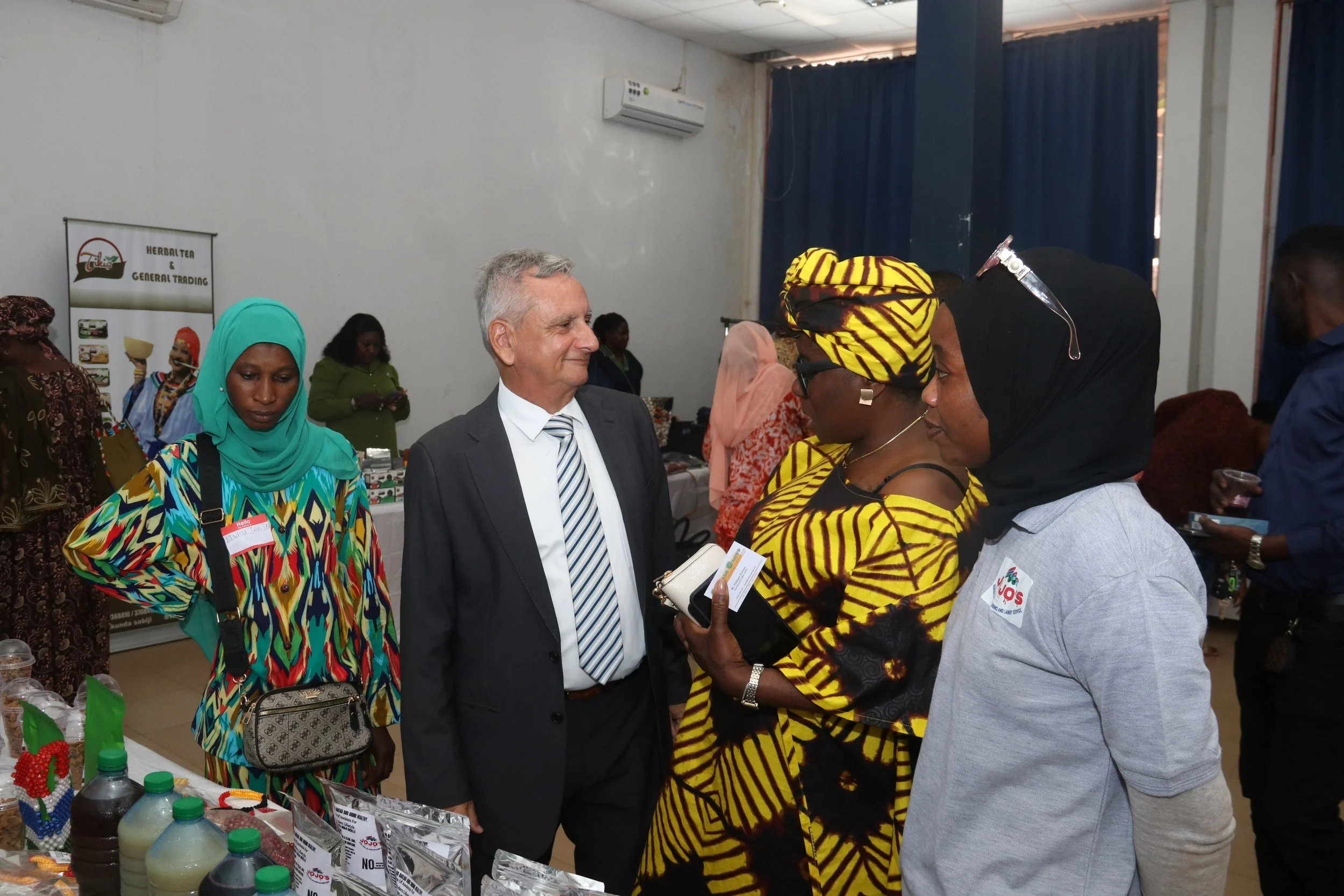 A diverse group of people including women in colorful clothing and a man in a suit are engaging in conversation at an indoor event. There are tables with products and a banner in the background that reads 'Herbal Tea & General Trading.'
