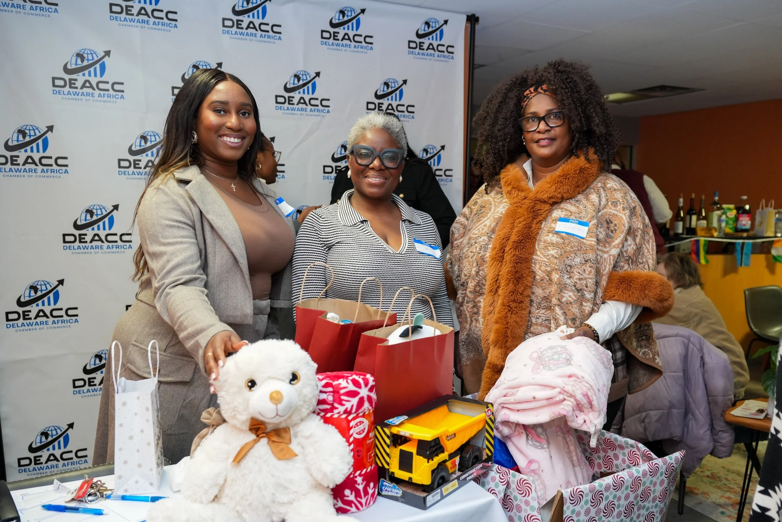 Three women standing together at a Delaware Africa Chamber of Commerce event, smiling at the camera. They are around a table with various gifts including a plush sheep, and there are gift bags and toys visible. The background features a DEACC backdro