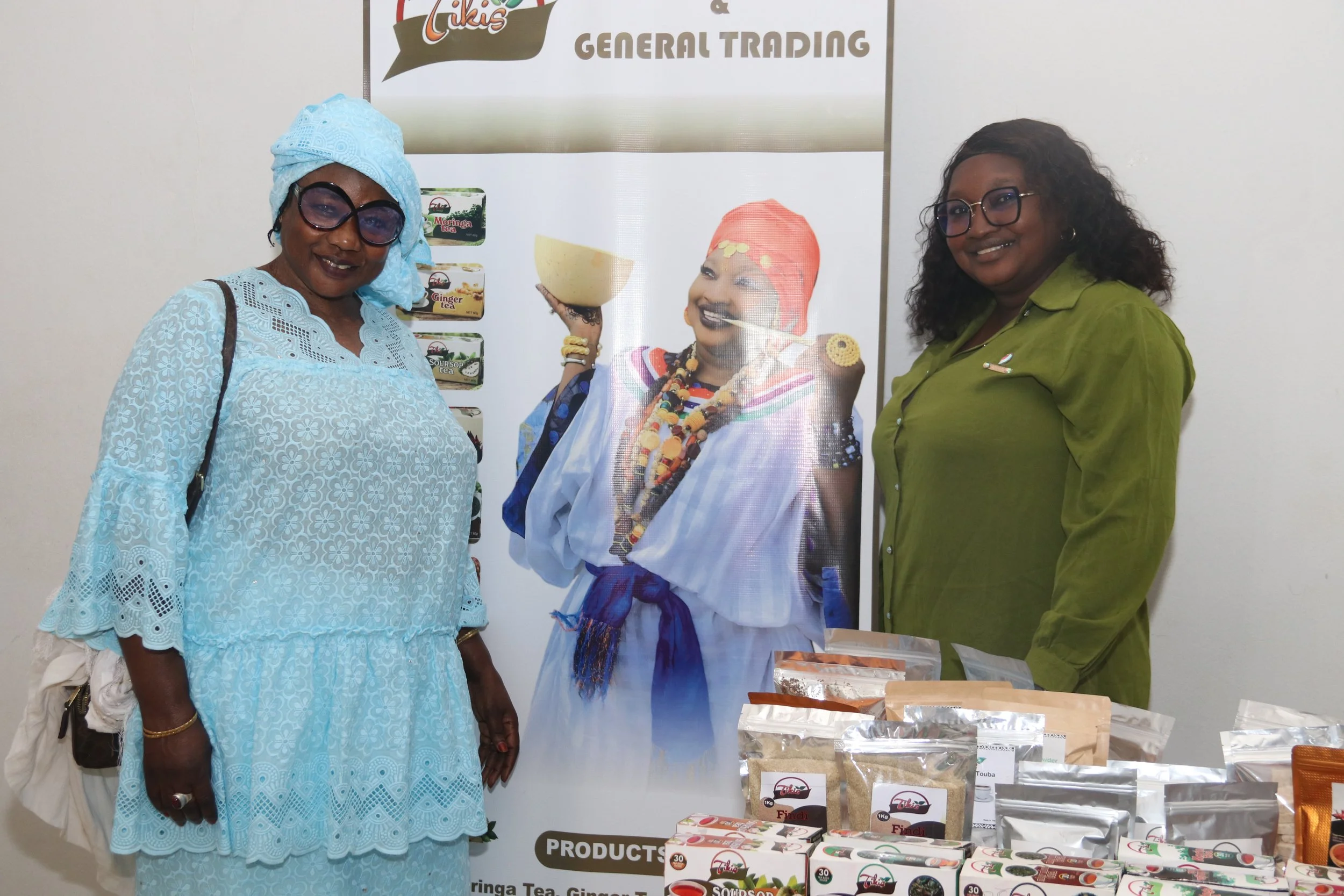 Two women standing next to a promotional poster for Likis, a brand of tea and flavorings, at a product exhibition. They are smiling and looking at the camera. The table in front of them displays various tea products and packaging.