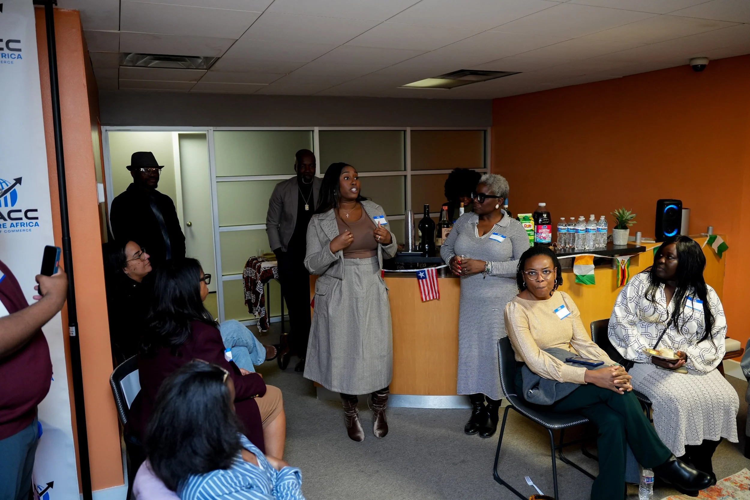 A diverse group of people attending a meeting or event in a room with orange walls. Some individuals are seated, while a woman is standing and speaking. There are drinks and supplies on a counter behind her, with a small plant and a speaker nearby. T