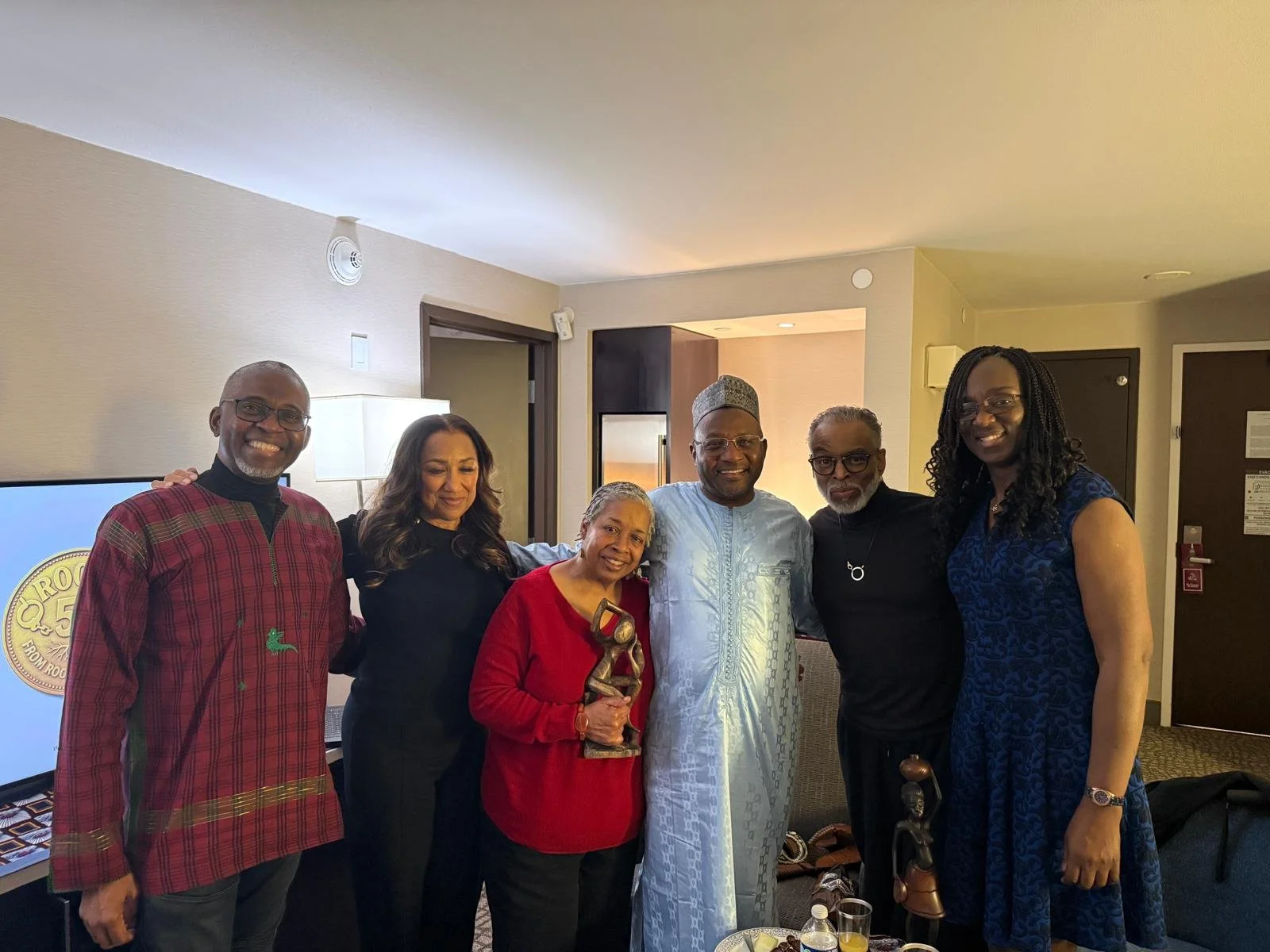Group of seven diverse people standing together in a hotel room, smiling for the camera, with one person holding a trophy or award.