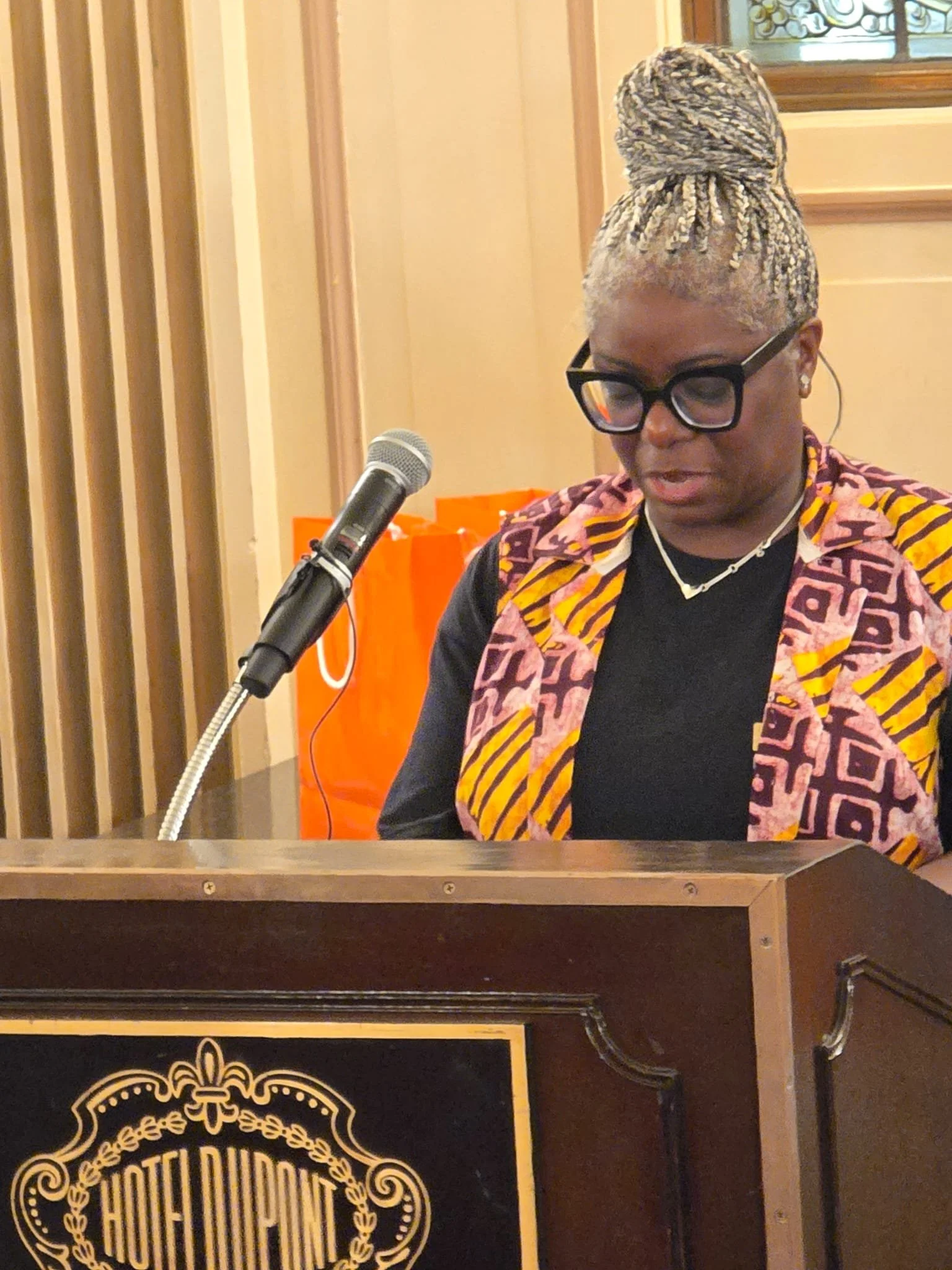 A woman with glasses and styled hair reading in front of a microphone at a podium with a Hotel du Palace sign.