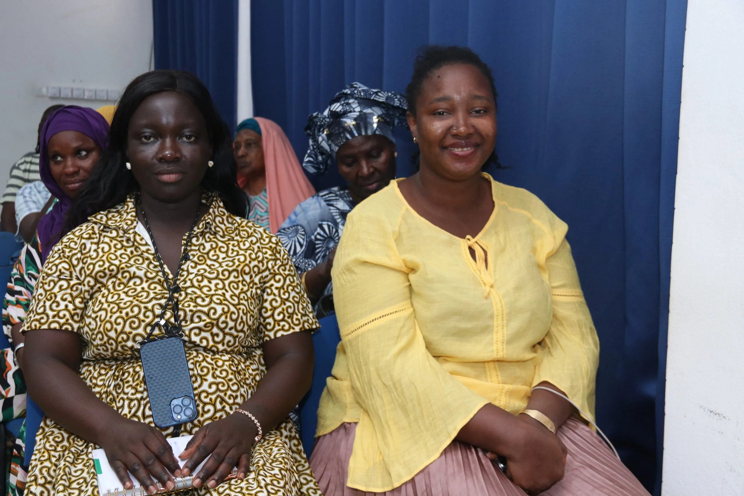 Group of women sitting in rows during an indoor event, with blue curtains in the background. Two women in the foreground are smiling; one wearing a yellow top and the other a patterned yellow dress, with one holding a smartphone and a notebook.