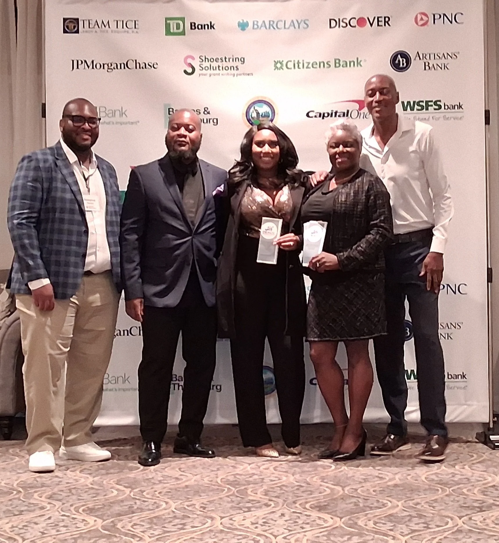 Group of five people standing together at an event with a backdrop showing various bank logos, including JPMorgan Chase, Citizens Bank, and PNC. Four of them are holding awards.