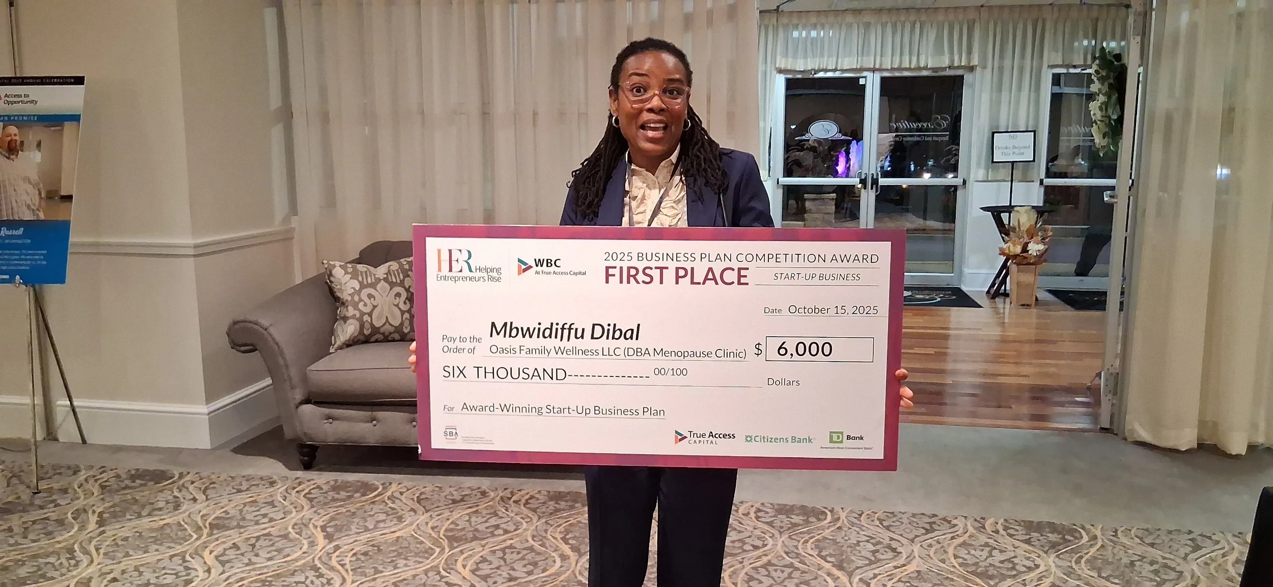 Woman with glasses holding large ceremonial check for $6,000 at an award event. Background includes a sofa, patterned carpet, and entrance to another room.