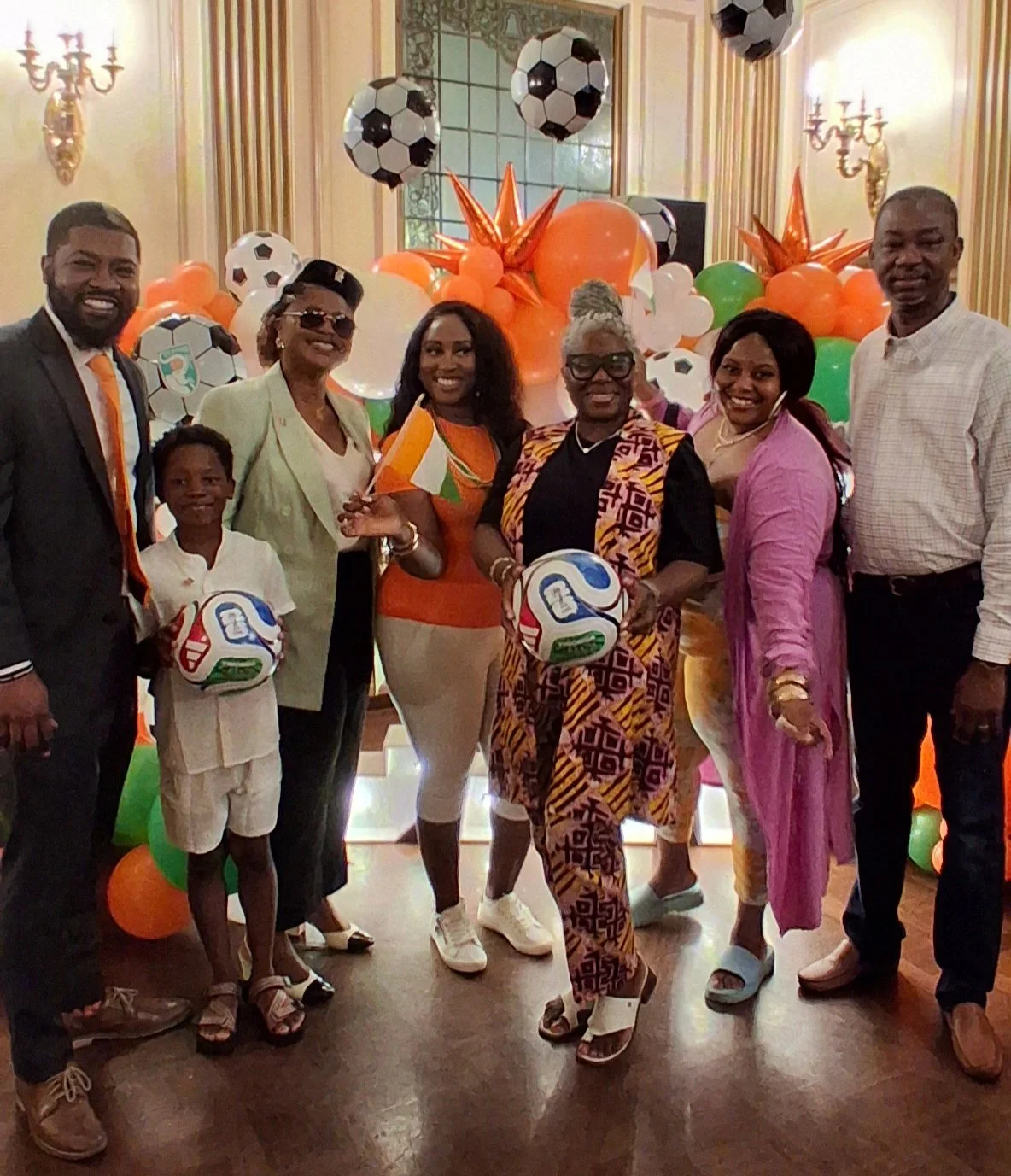 A group of seven people, including a child, standing together at a celebration with orange, green, and white balloons, and soccer ball decorations in the background.
