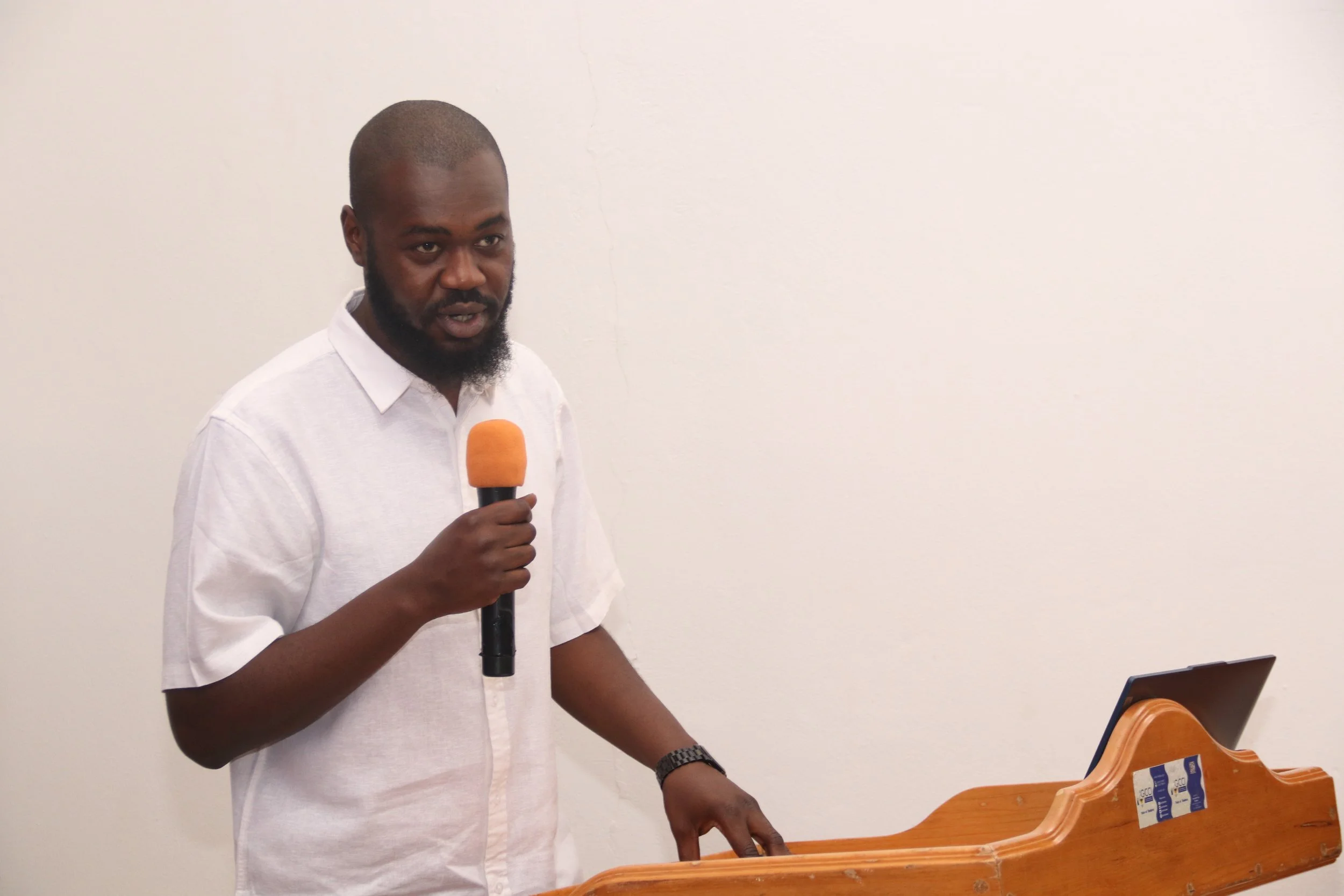 A man with a beard and short hair wearing a white shirt holding an orange microphone and standing next to a wooden music stand with a laptop on it, in front of a plain white wall.
