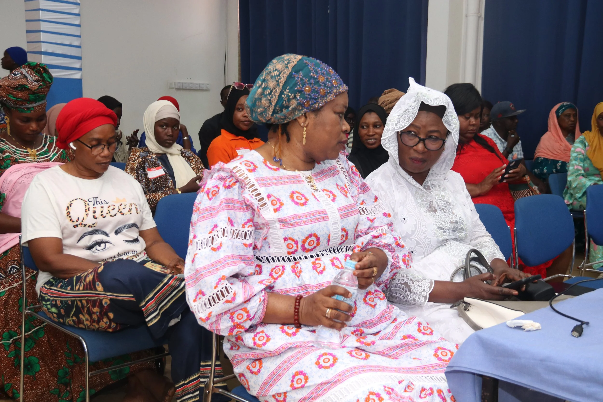 Several women, many wearing traditional clothing and head coverings, are seated in a room with blue curtains, some using their phones, with a few looking ahead or down.
