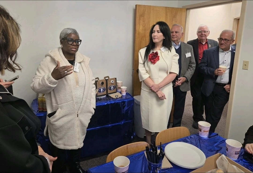 Group of six people standing around a table with disposable cups and plates in a meeting room. One woman in a white coat is speaking, while the others listen. There are beverage containers on a table in the background.