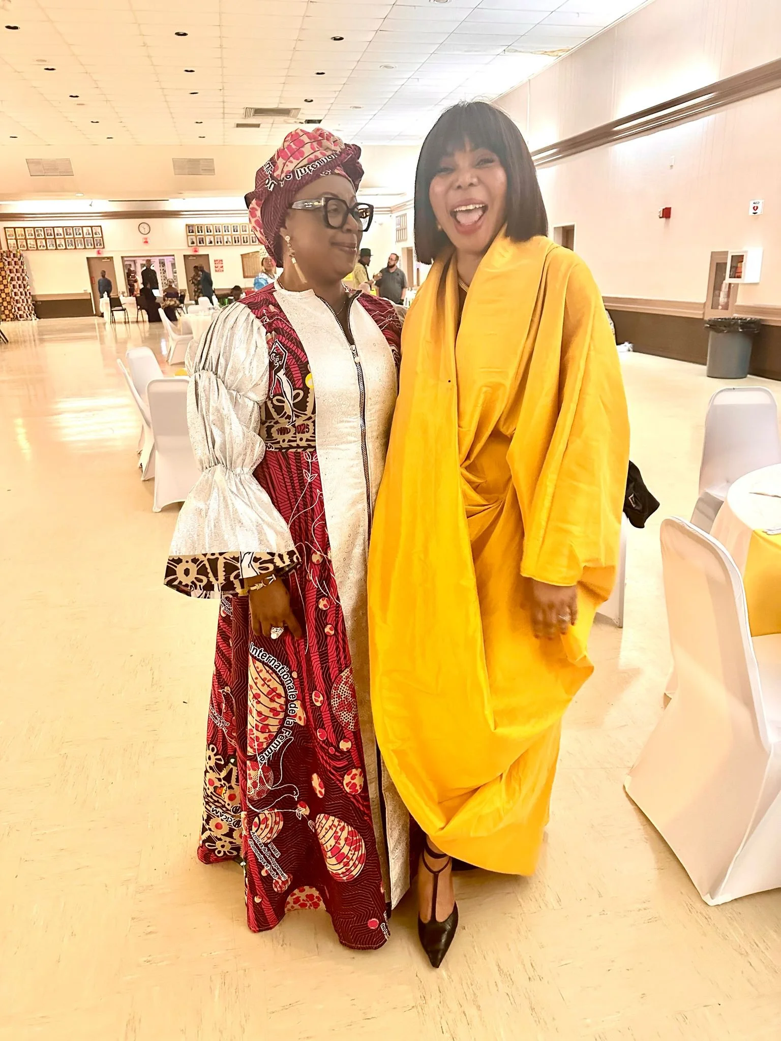 Two women standing closely together indoors, smiling and enjoying the moment. One woman is dressed in traditional African attire with a matching headwrap, glasses, and earrings. The other woman is wearing a bright yellow outfit with a wide smile. The