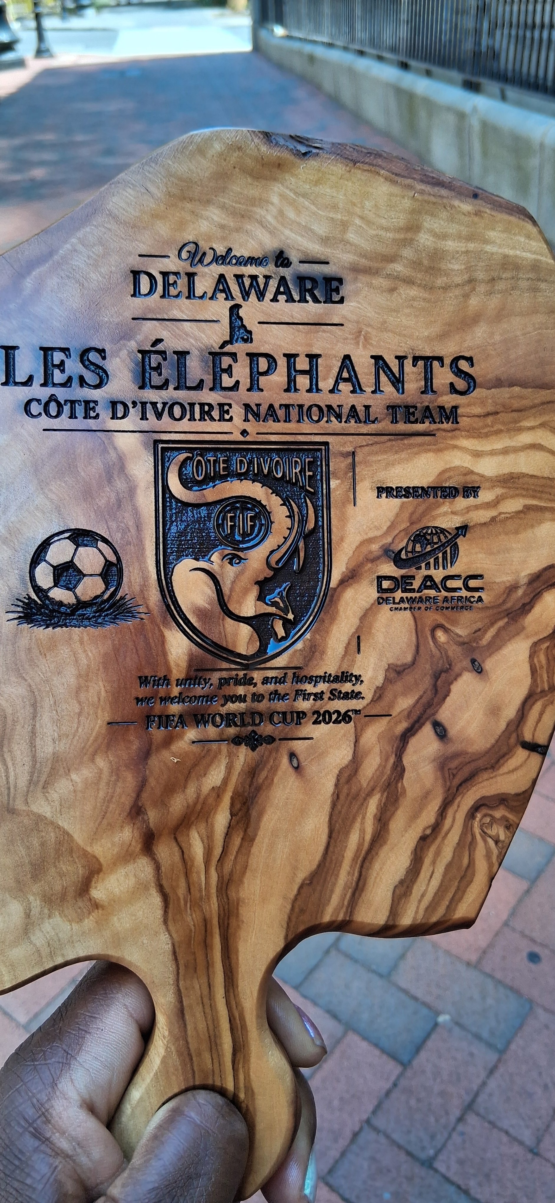 A wooden plaque with inscriptions and logos welcoming to Delaware and the Ivory Coast National Team, featuring the FIFA World Cup 2026 logo and a soccer ball, presented by DEAC Delaware Africa.