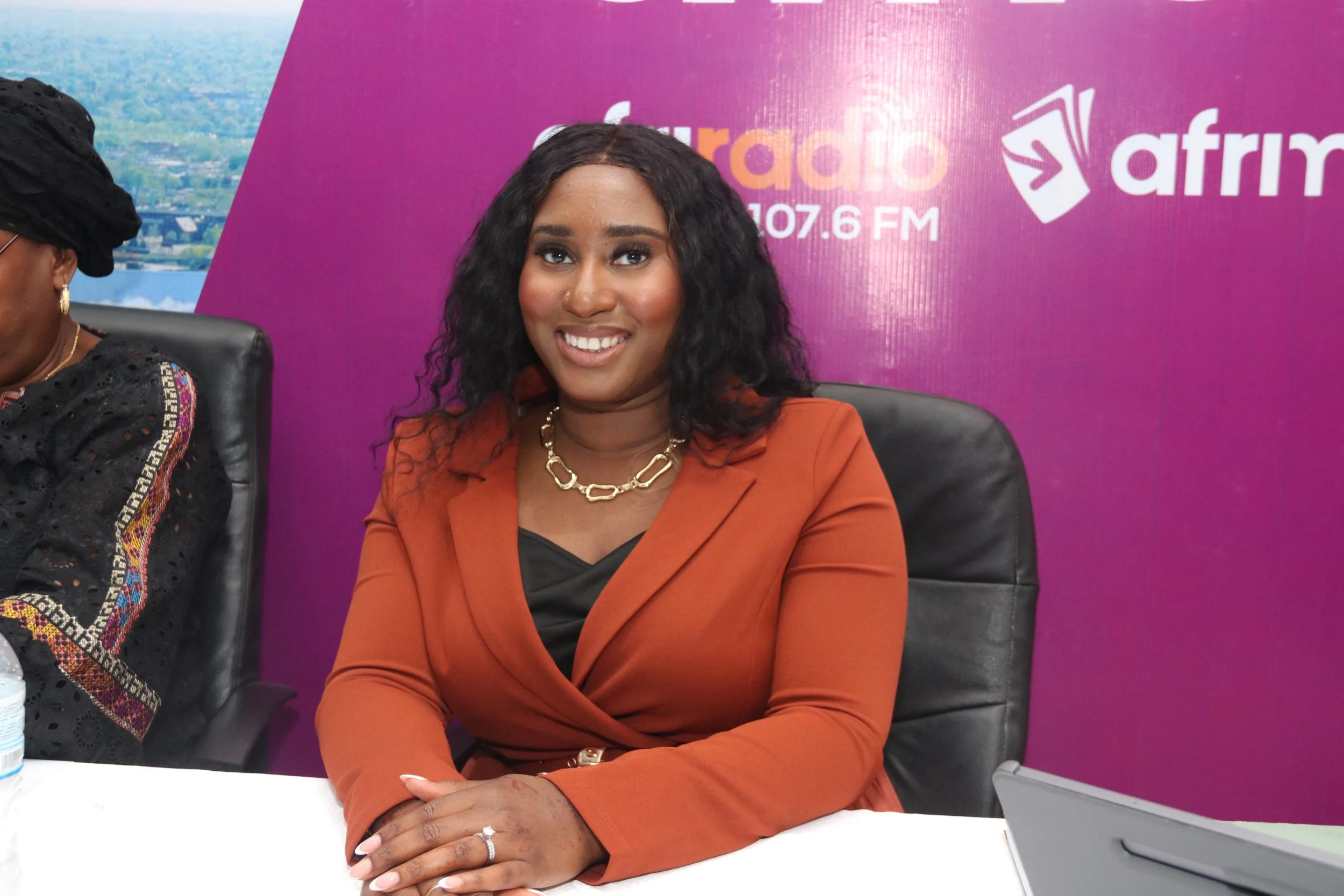A woman with dark curly hair, wearing an orange blazer, gold necklace, and jewelry sitting at a table with a purple background featuring logos of a radio station and a company.