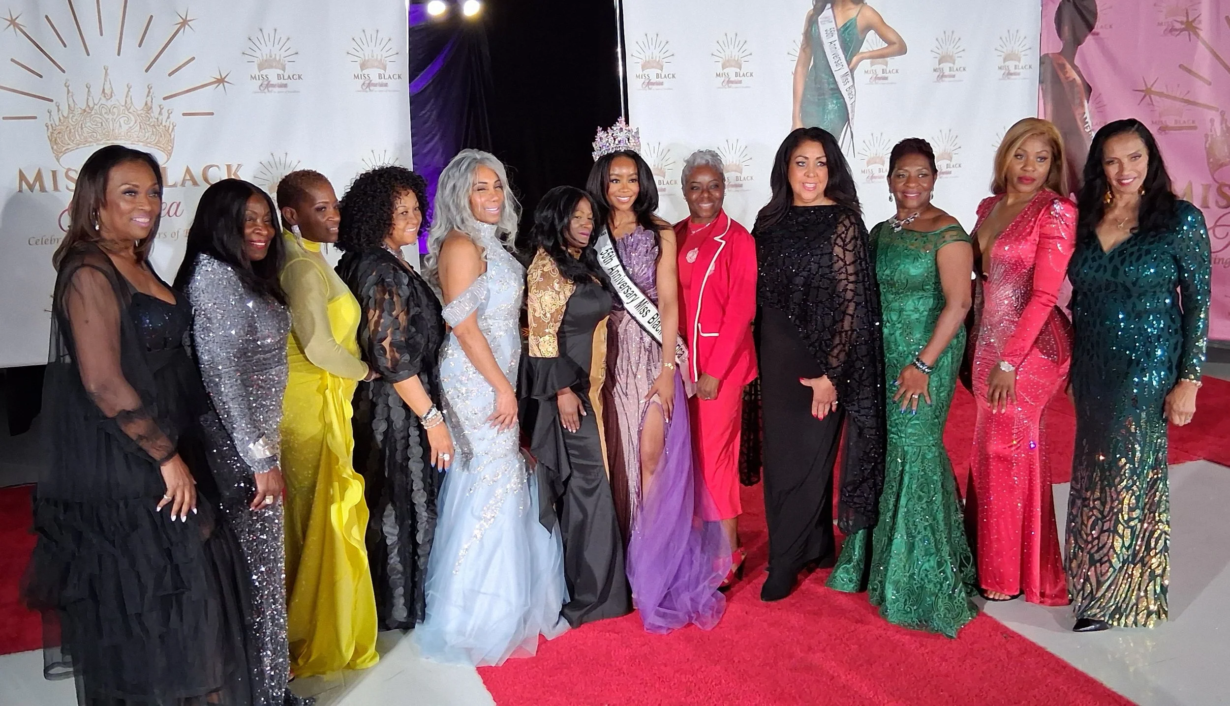 A group of women in formal evening gowns and dresses standing on a red carpet at an event. There is a woman in the center wearing a crown and sash indicating she is Miss Black Universe. The background features a step-and-repeat banner with the Miss B