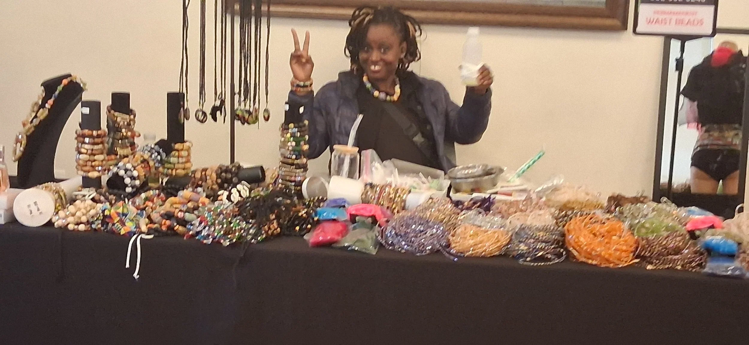 Woman smiling behind a table filled with colorful beaded jewelry and accessories, making a peace sign with her fingers.
