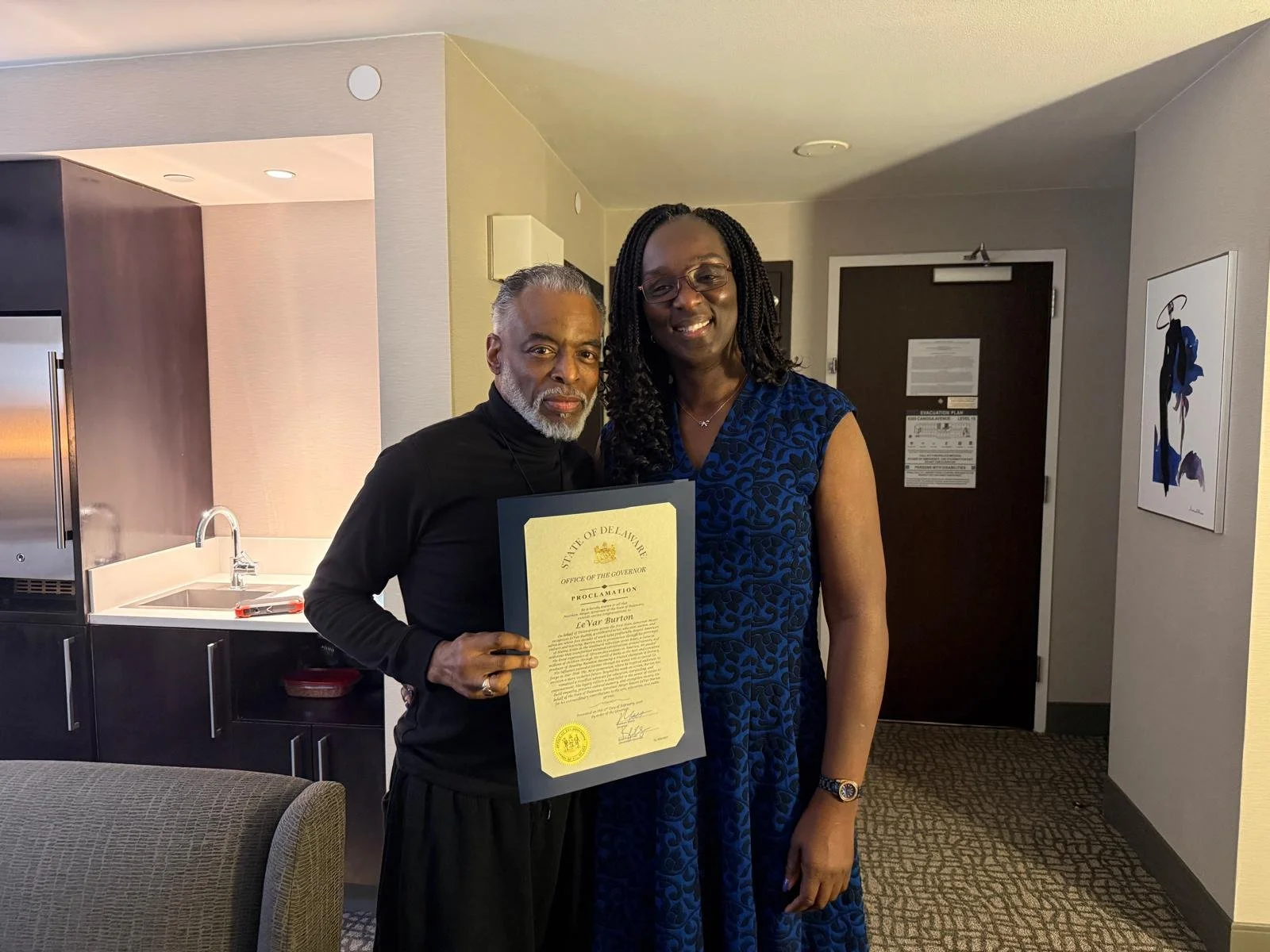 A man and woman standing together indoors, the man holding a proclamation document, both smiling at the camera.