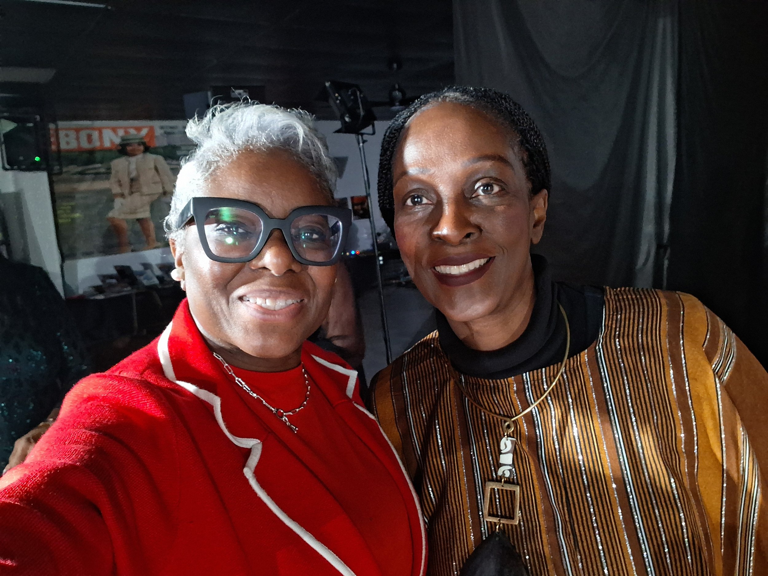 Two women taking a close-up selfie at an event, with one woman wearing glasses and a red top, and the other woman wearing a striped jacket and possibly a necklace.