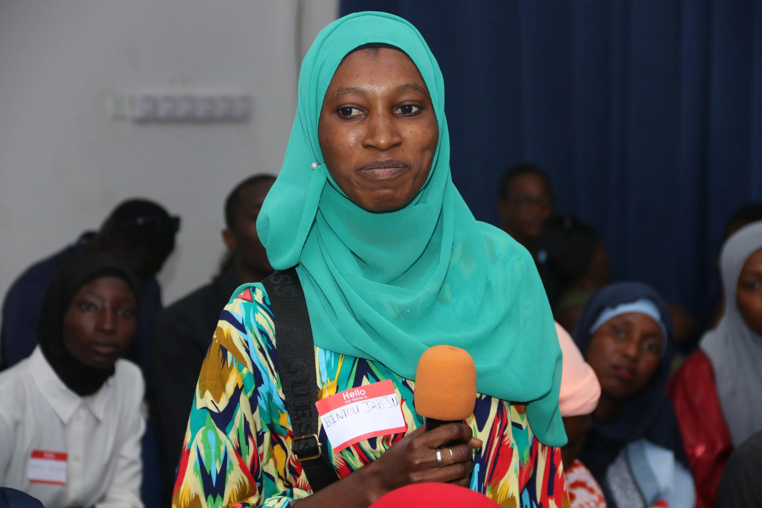 A woman wearing a turquoise headscarf and colorful dress speaking into an orange microphone at a conference or meeting. She has a name tag that reads 'Bintou Jarr.' Several people are seated in the background, some with headscarves.