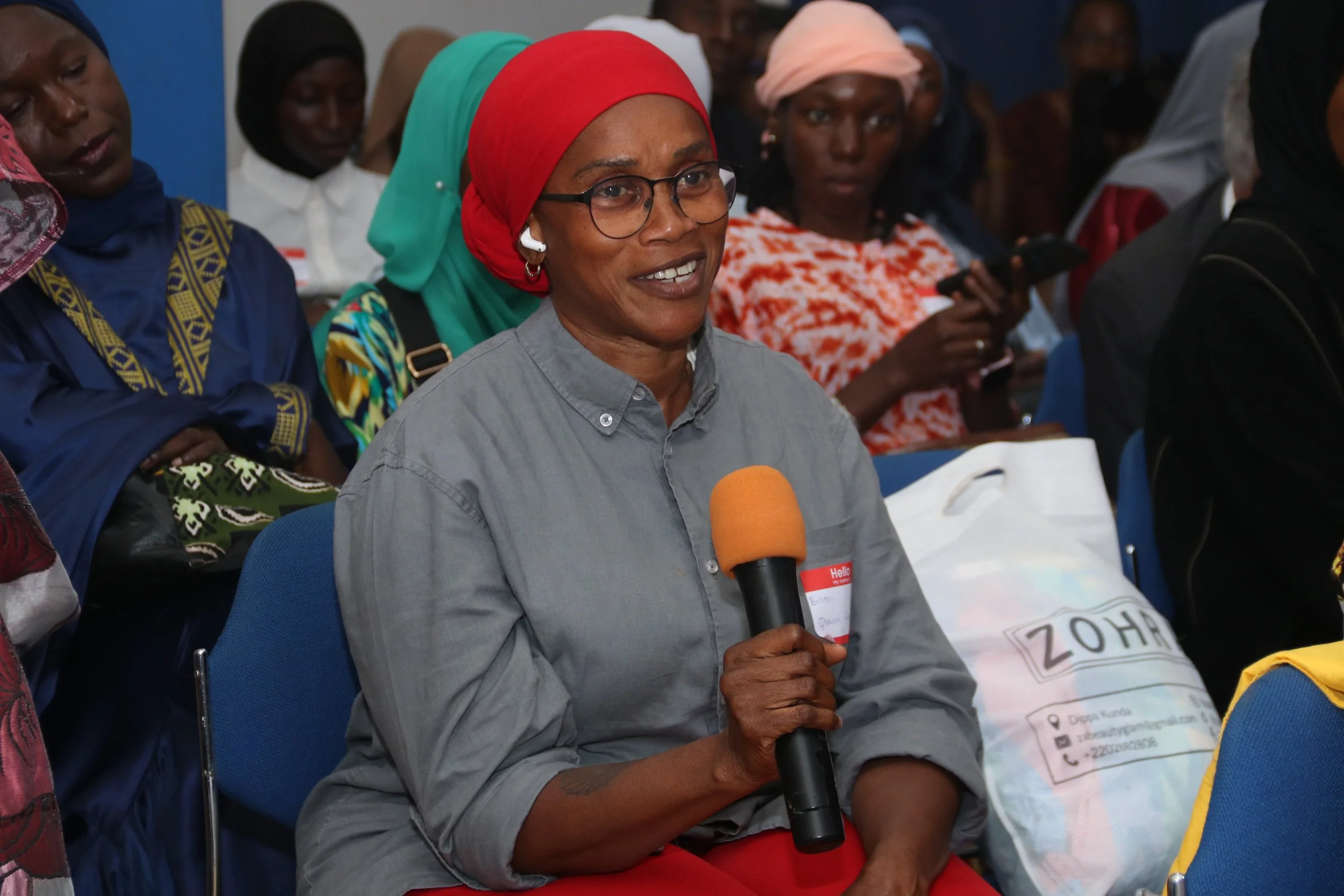A woman wearing glasses and a red headscarf speaking into a microphone at a gathering or conference, surrounded by other attendees.