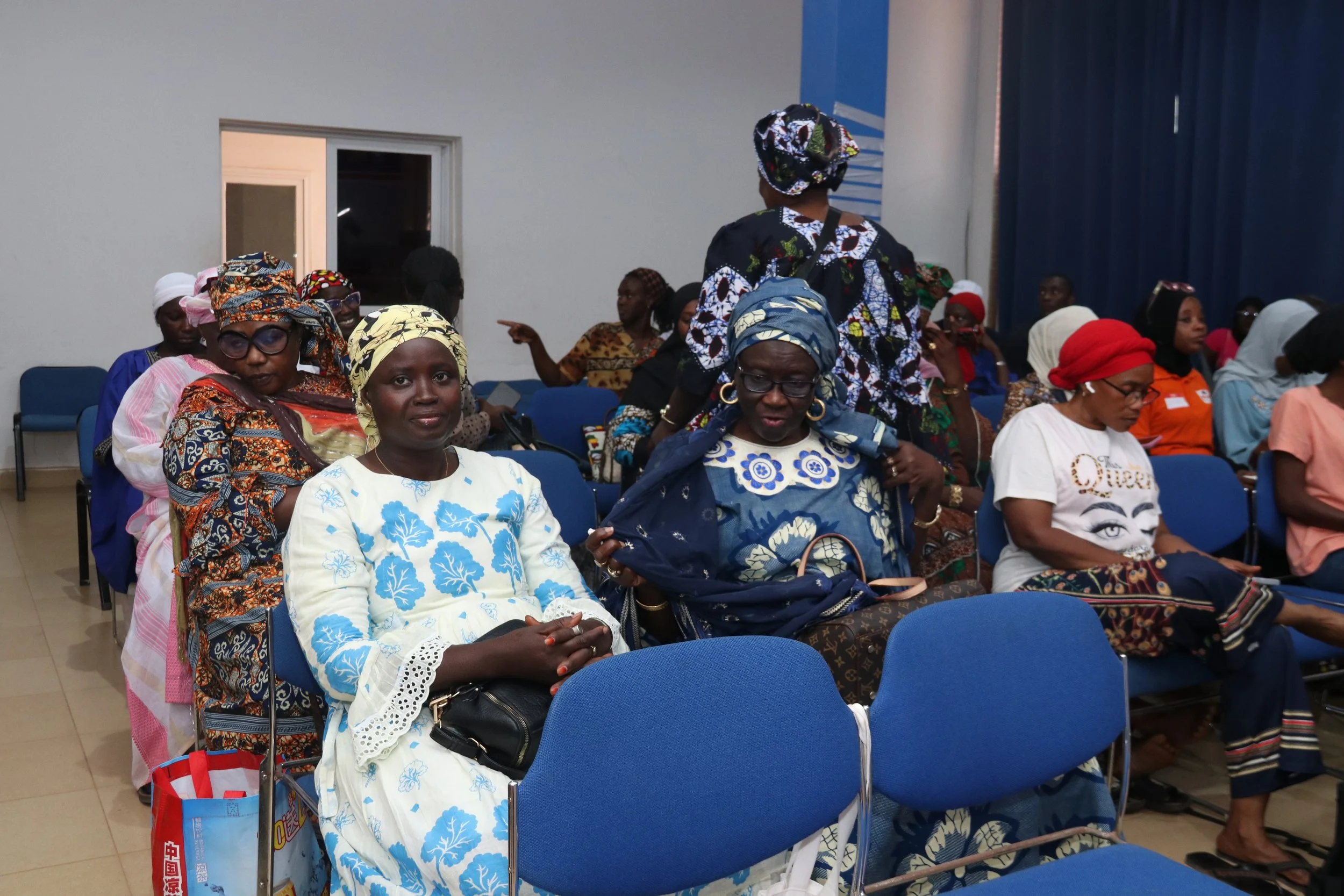Group of women attending a meeting or event in a room with blue chairs, wearing colorful traditional clothing and head coverings.