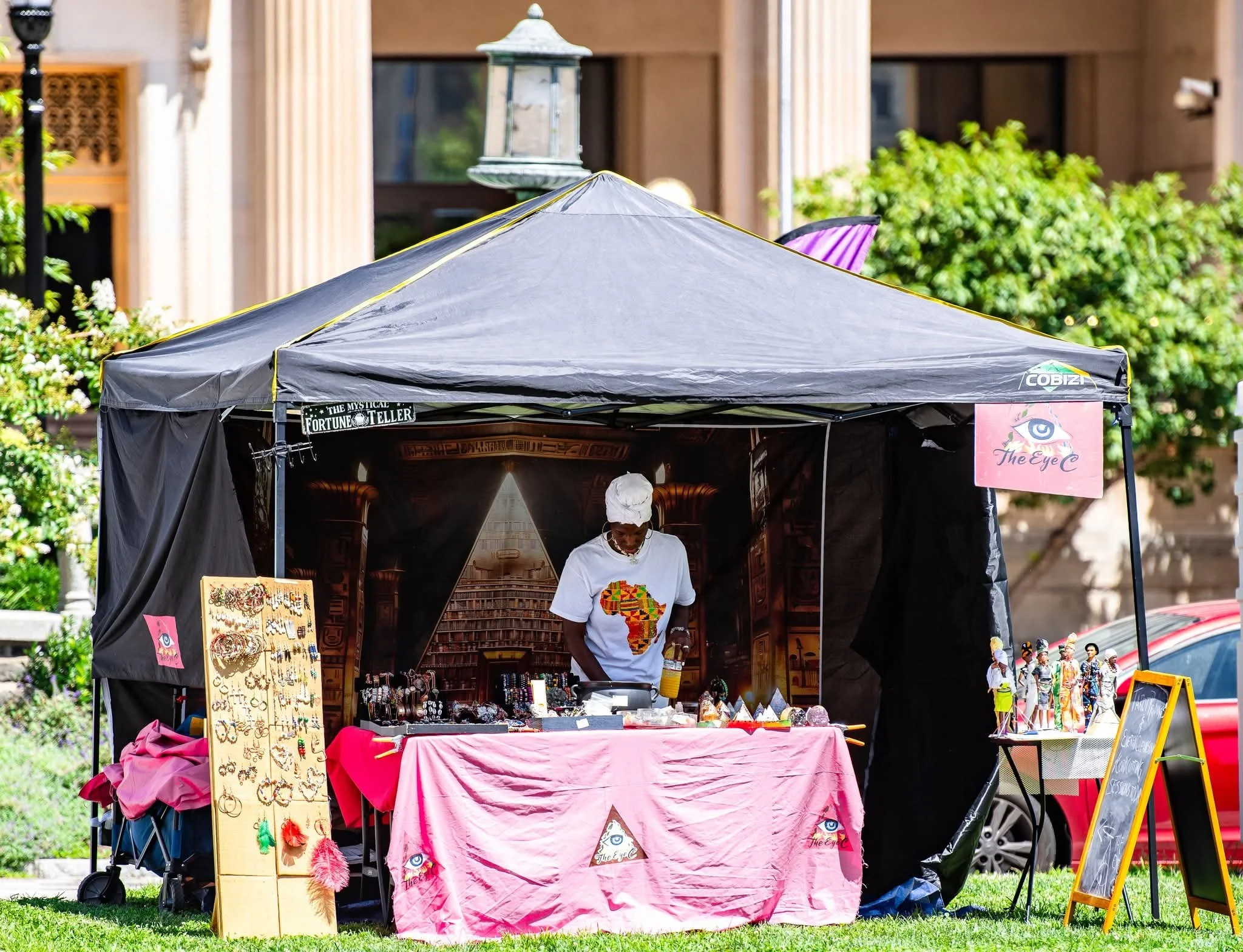Outdoor market stall with a pink cloth covering and a sign titled "The Eye" displaying jewelry and accessories, with a person wearing a white shirt and head wrap inside.