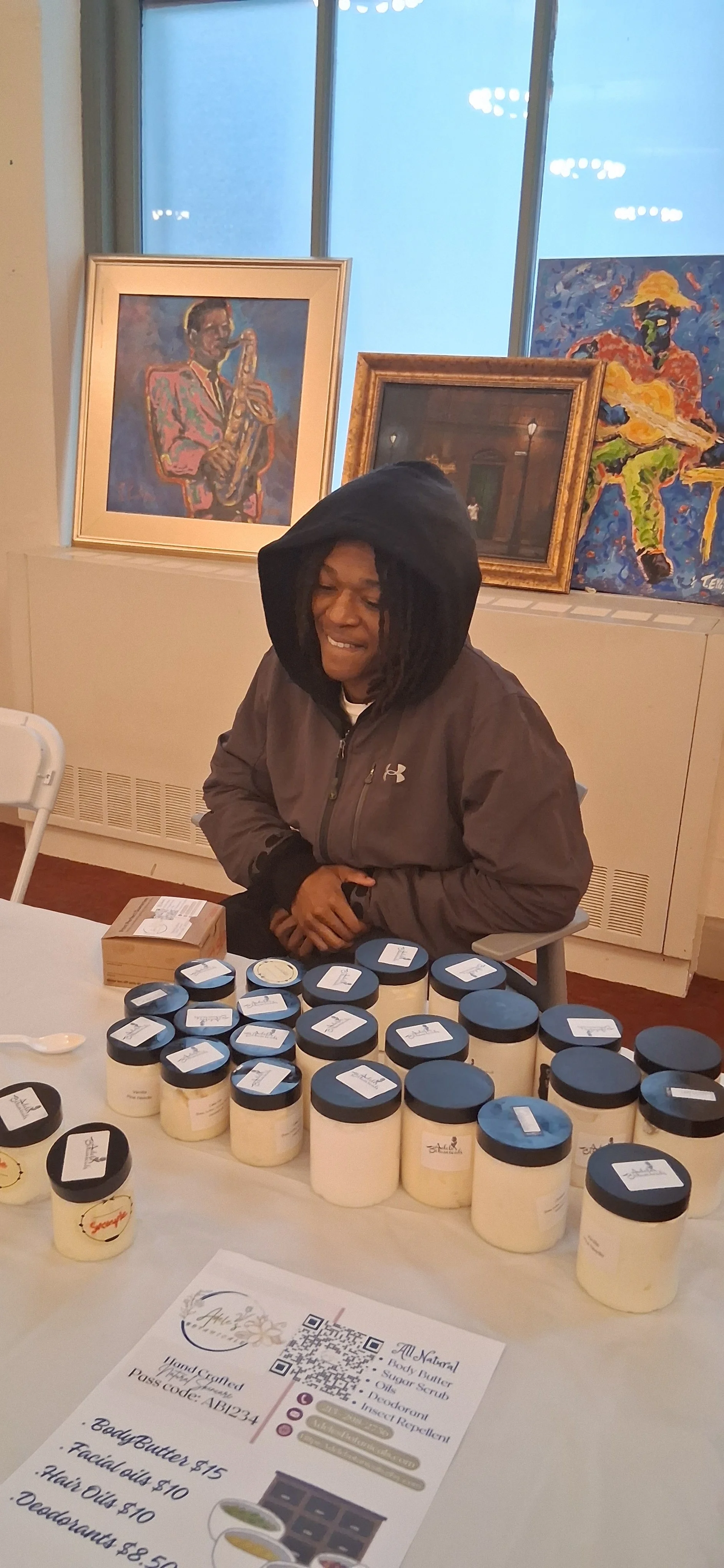 Woman sitting at a table with jars of body butter for sale, in an art gallery with paintings on the wall behind her.