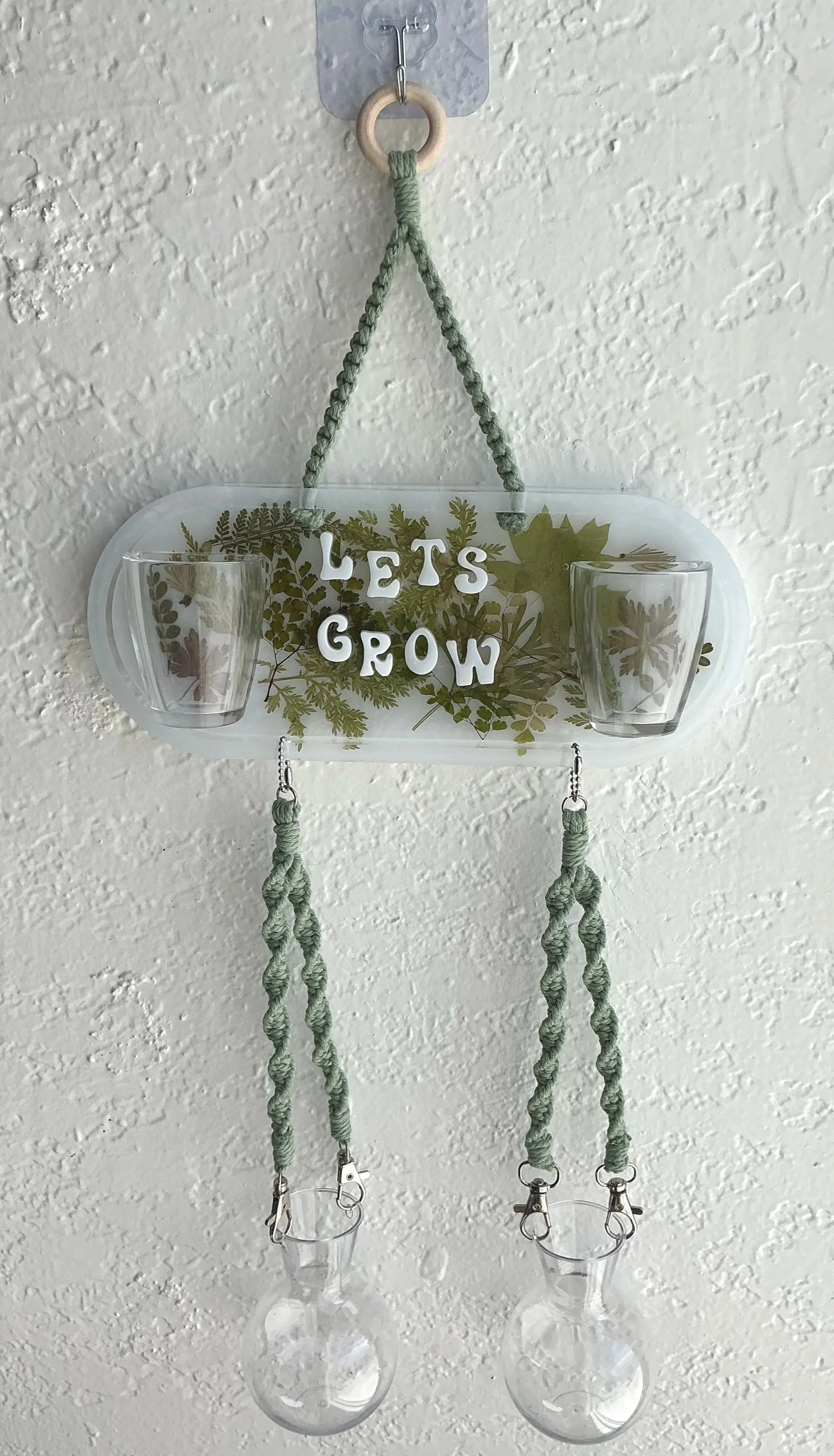 Full front view of the actual propagation station without plants, showing the wooden hanging ring, pressed botanical resin panel, four empty vessels, sage macramé, and silver hardware.