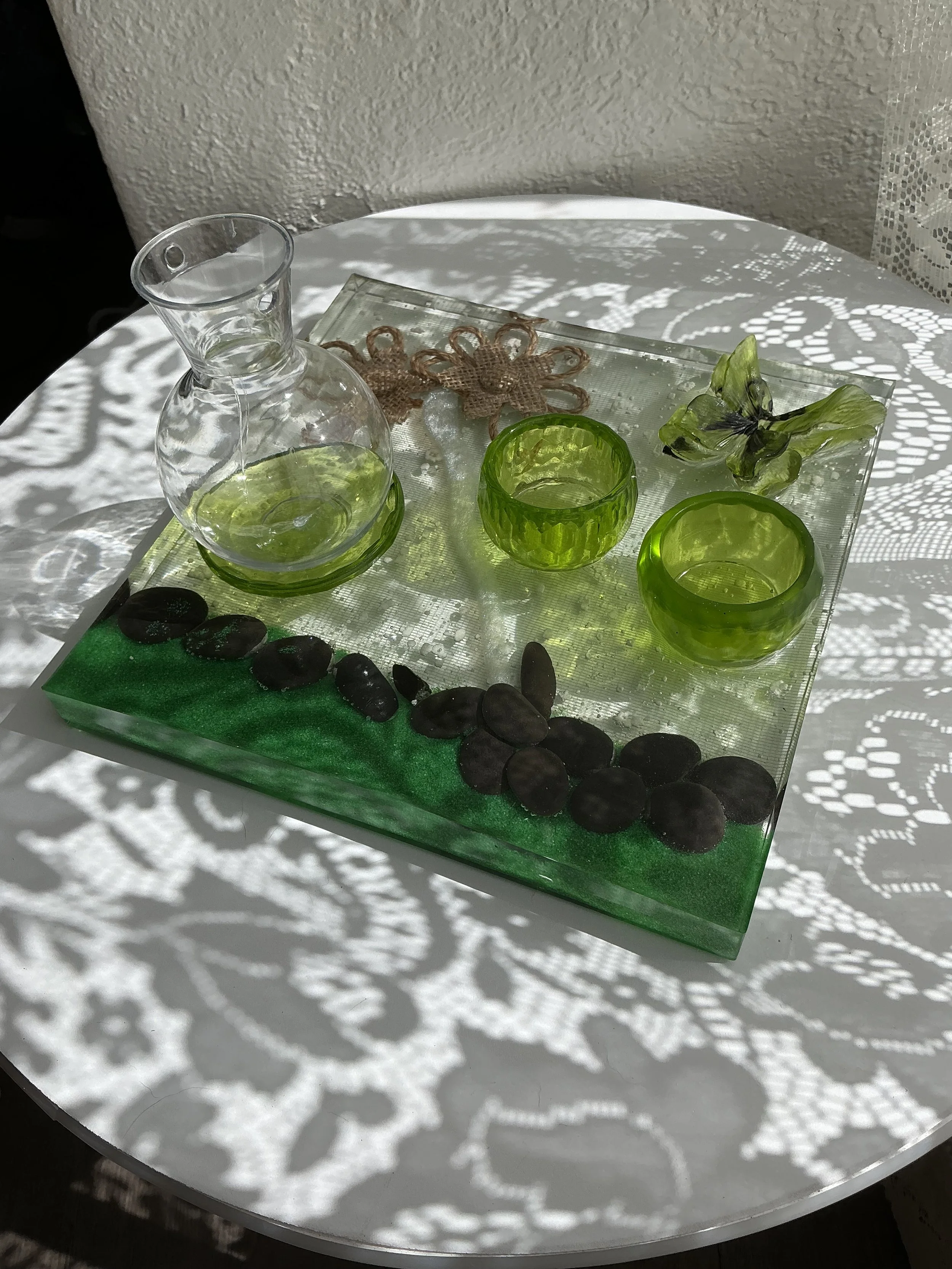 Low side view revealing the thickness of the clear resin base, shimmering emerald green edge and embedded black stones. A glass propagation vase and two bright green plant holders rise above the surfa