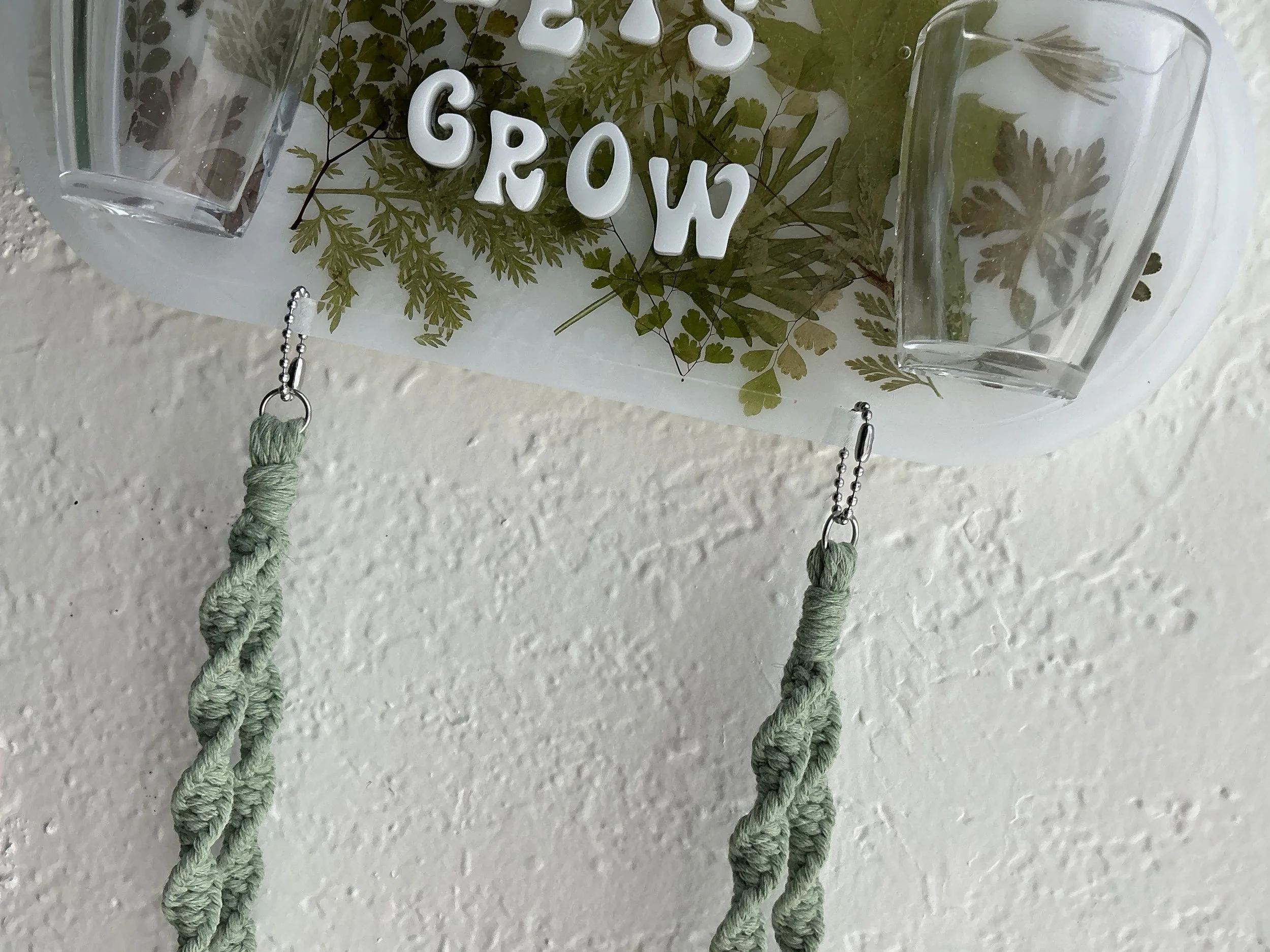 Angled close-up of the resin panel’s lower edge, silver ball-chain connections, and sage macramé hangers that support the removable propagation vases.
