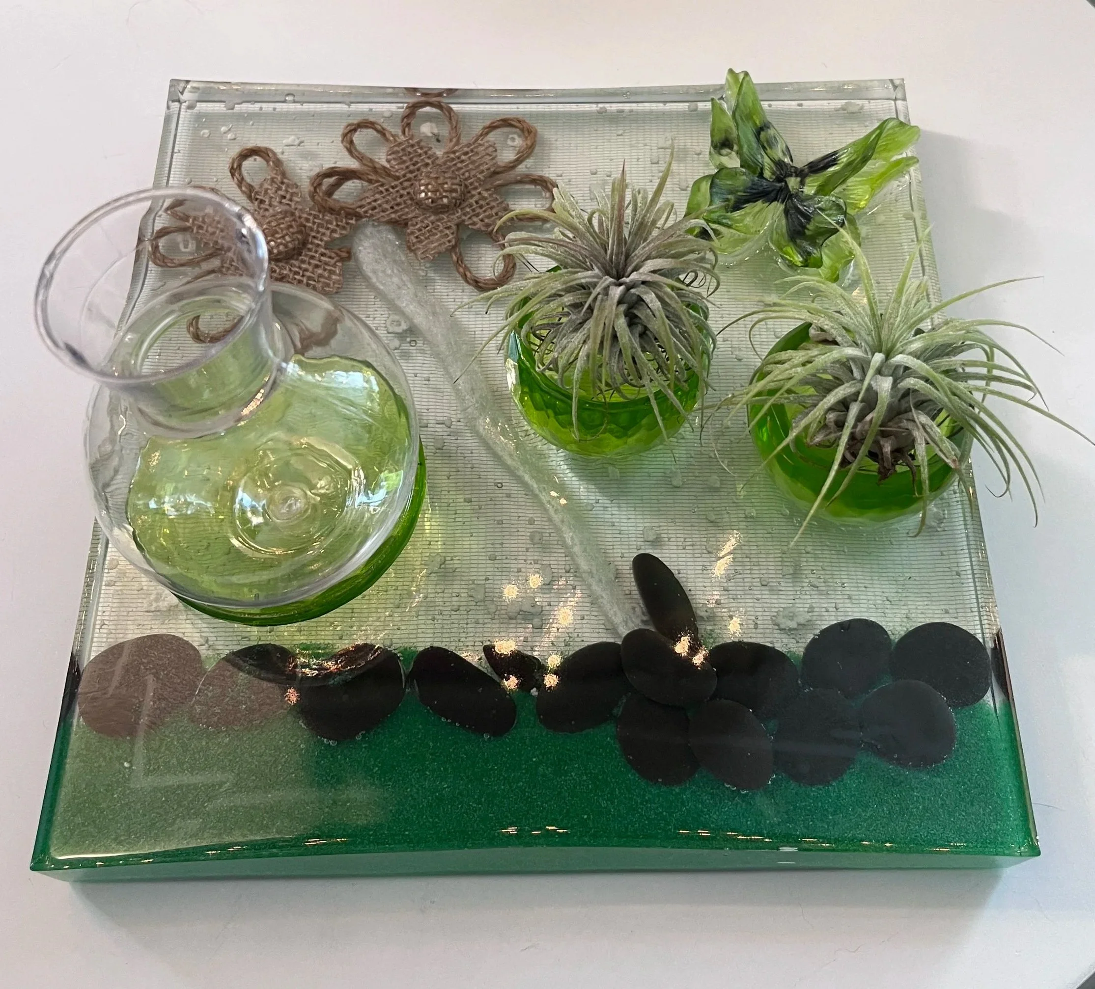 Angled view of the finished resin propagation station with two air plants in green holders and a glass vase for cuttings. Black stones, emerald resin, burlap flowers and a butterfly create a tiny gard