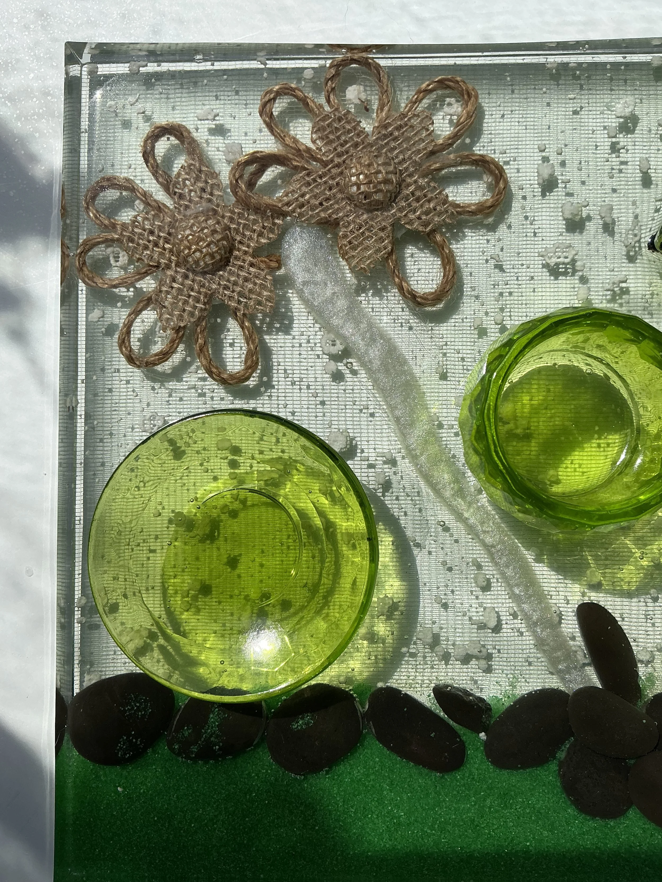Close-up of two rustic burlap flowers and a shimmering white branch embedded in clear resin. A lime-green plant holder and black stone border add color and texture to the botanical design.