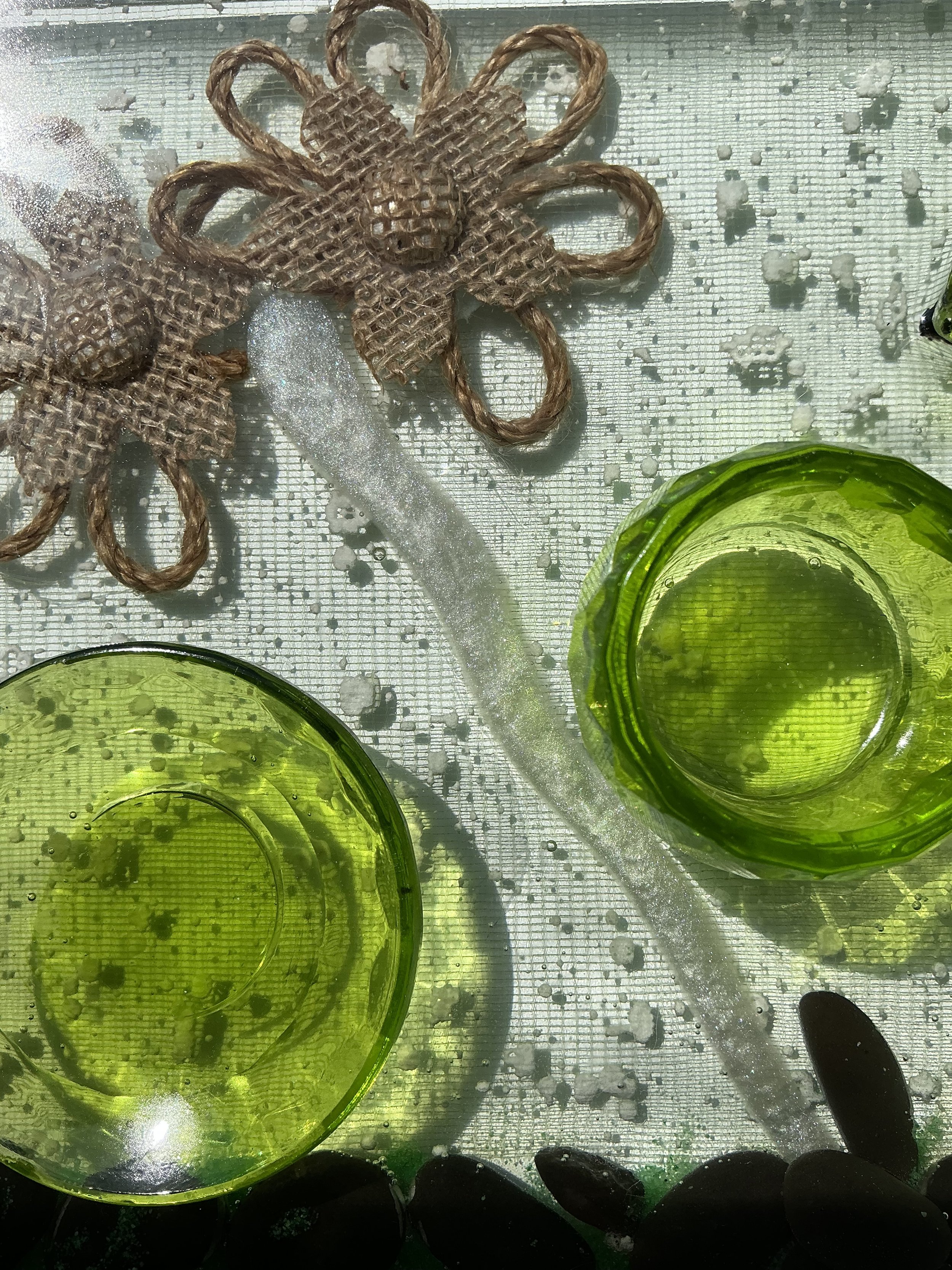 Close-up of two rustic burlap flowers and a shimmering white branch embedded in clear resin. A lime-green plant holder and black stone border add color and texture to the botanical design.