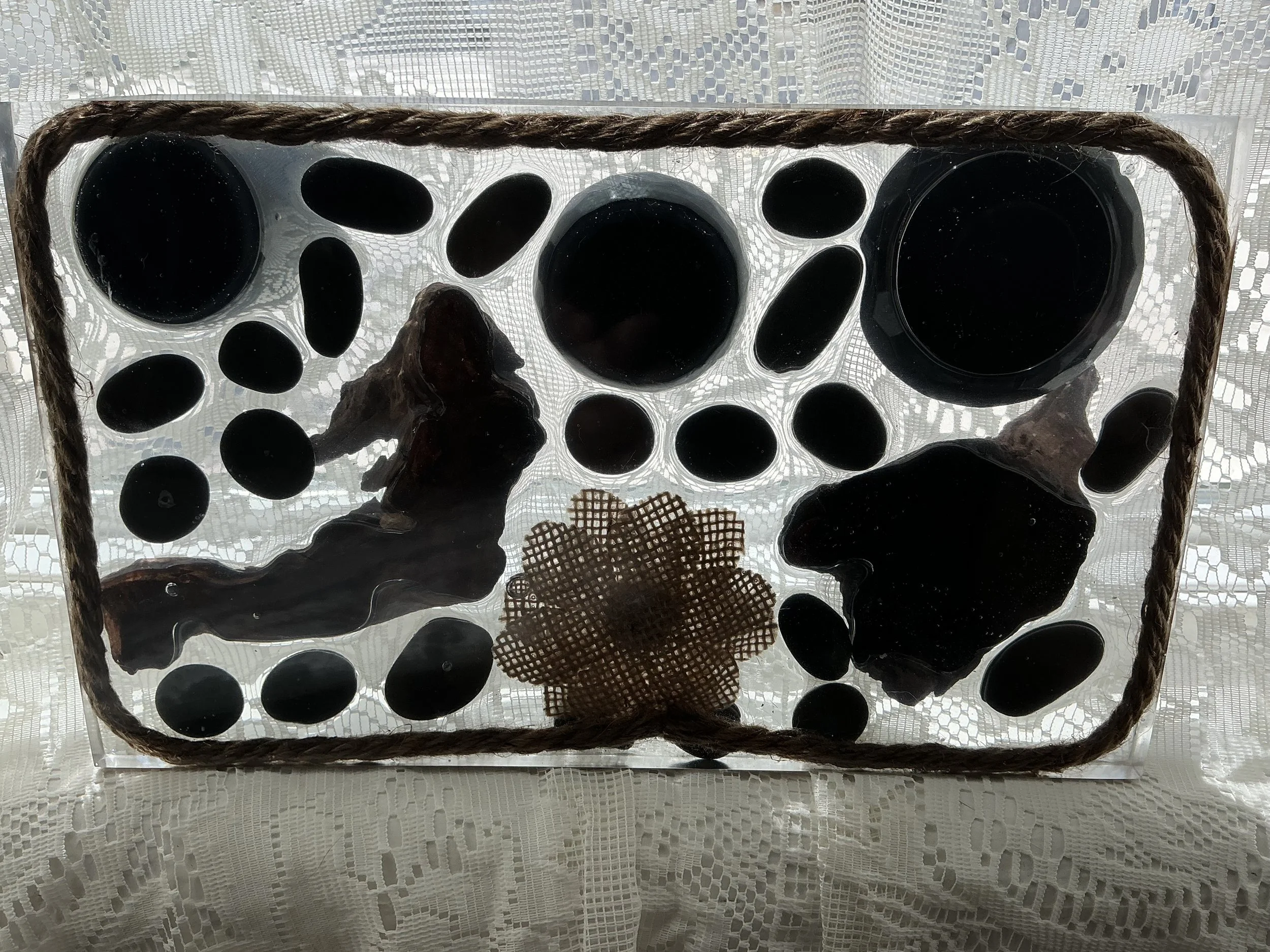 Backlit underside view through the clear resin station showing silhouettes of embedded black stones, driftwood, jute border and burlap flower against a lace curtain, highlighting the resin's transpare