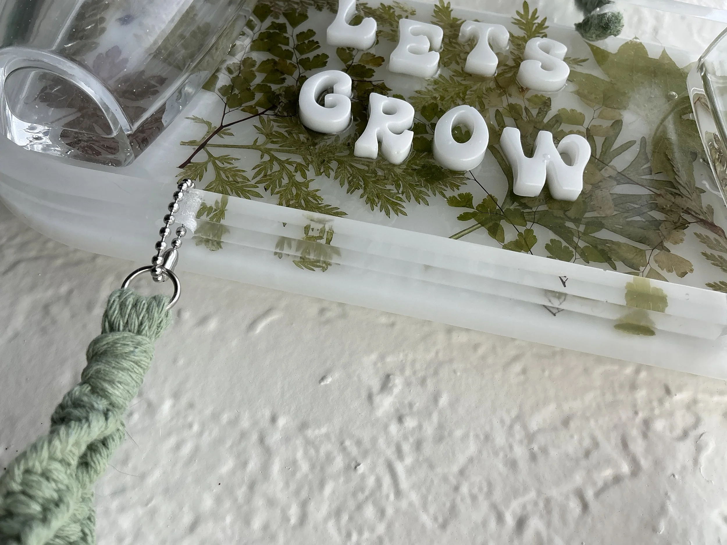 Detailed angled view of the raised white lettering, real pressed foliage inside the clear resin, layered panel edges, and one lower ball-chain attachment point.